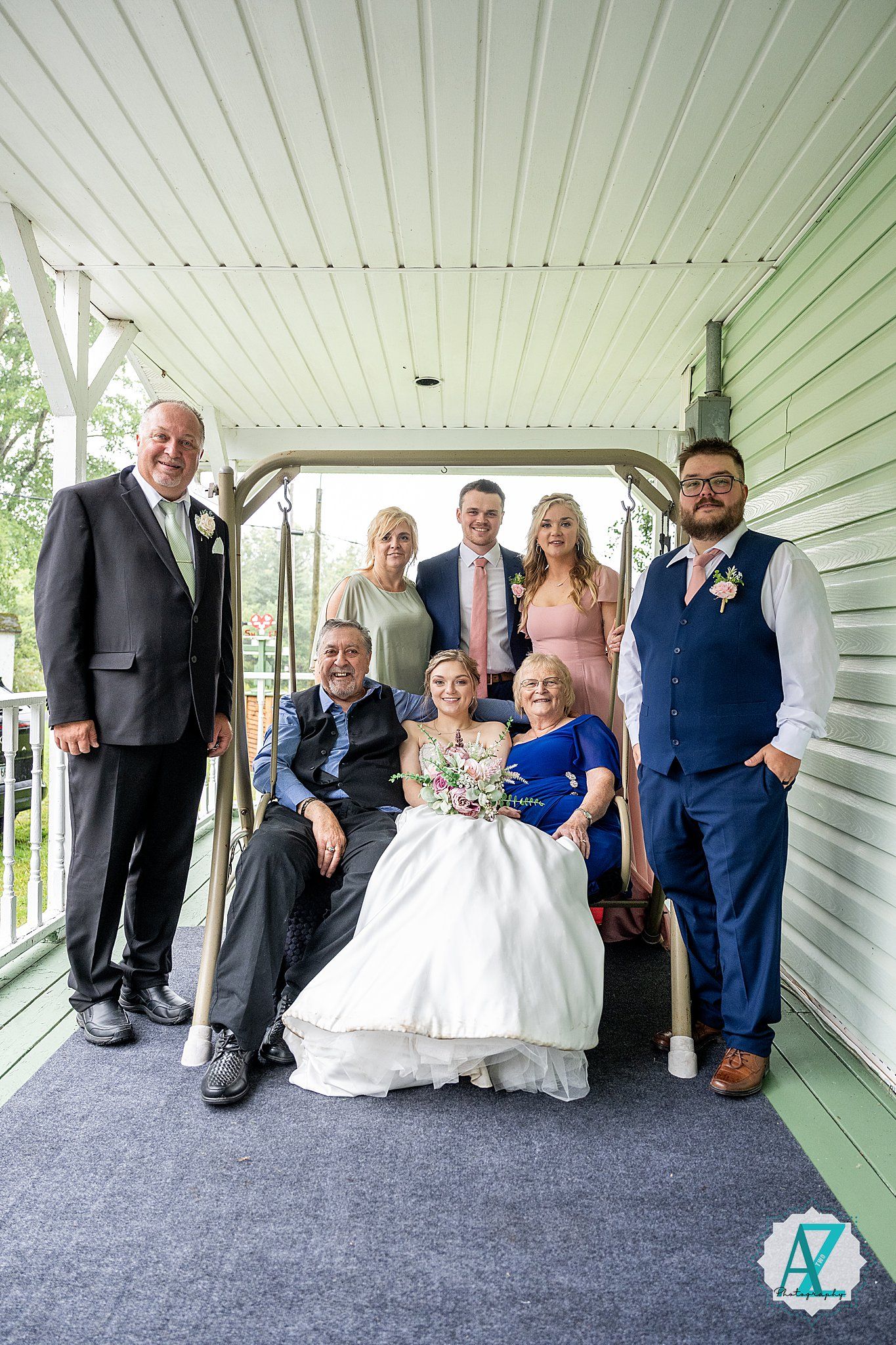 Family and wedding party members gather for formal photos on a covered porch with blue carpeting.