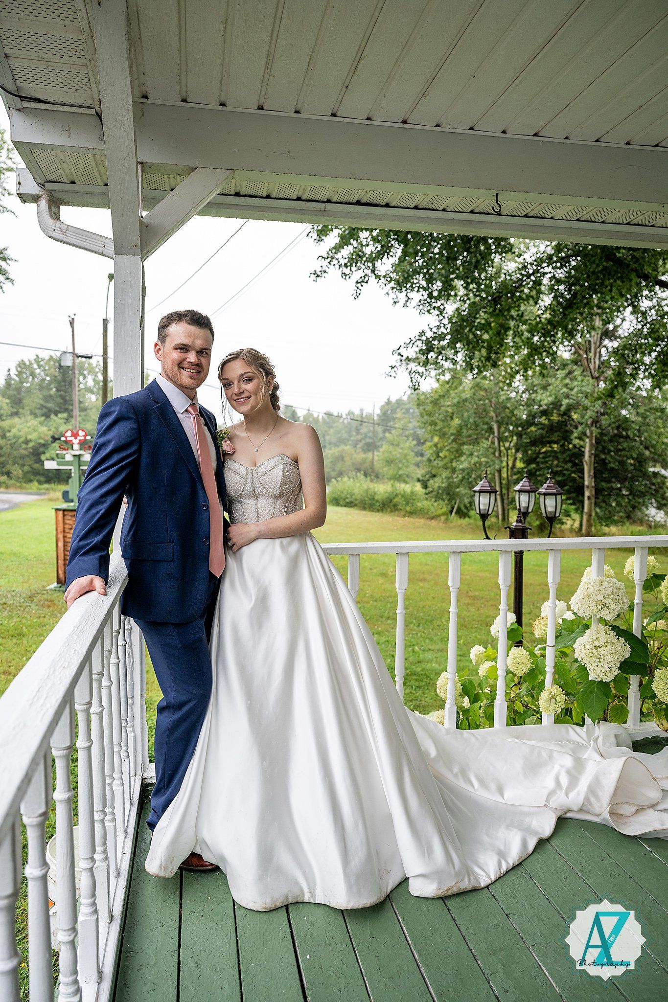 A newlywed couple stands on a white porch with decorative railings and hydrangea flowers in the background.
