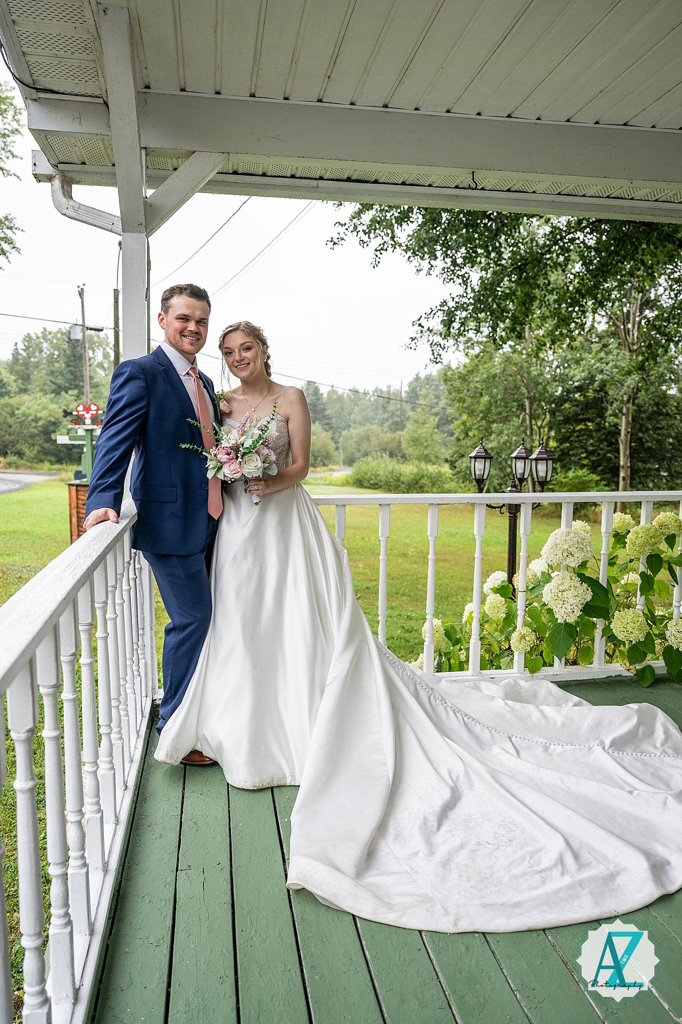 A couple poses on a white farmhouse porch with spindle railings and green floorboards during their wedding photoshoot.
