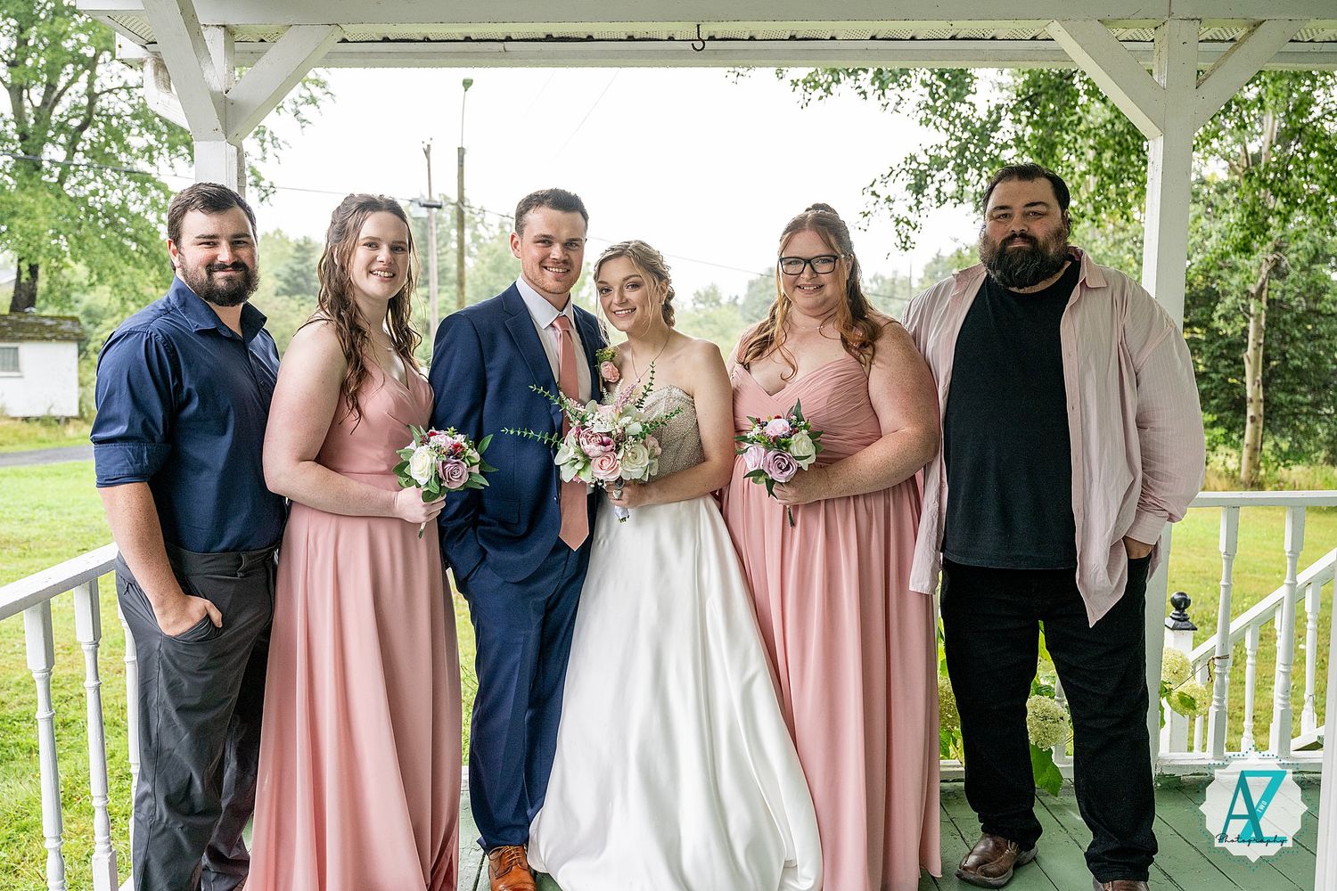 Wedding party in blush pink dresses and navy suits pose together on a white gazebo on a sunny day.