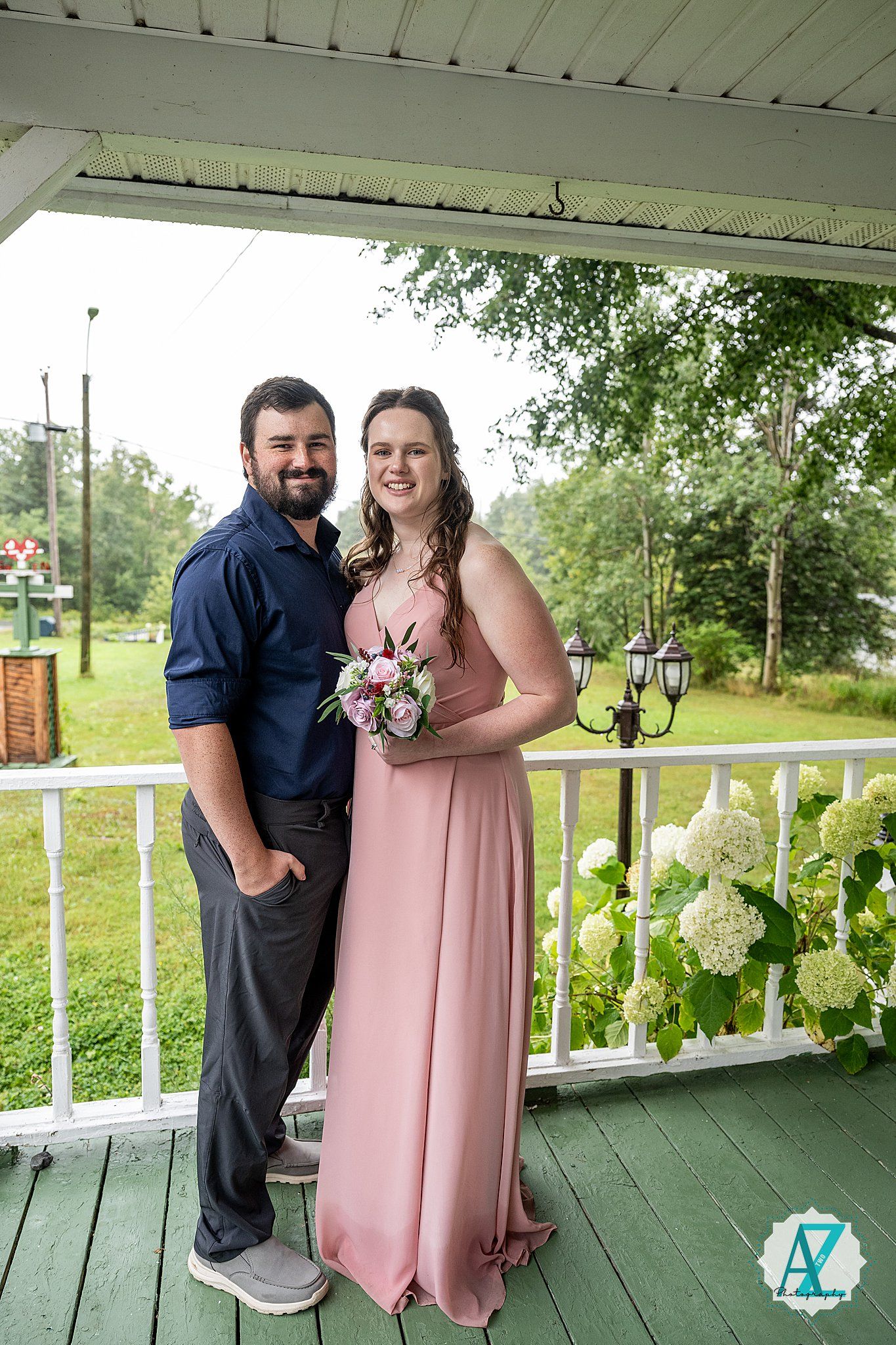 Wedding couple standing on a covered white porch with lush greenery in the background.