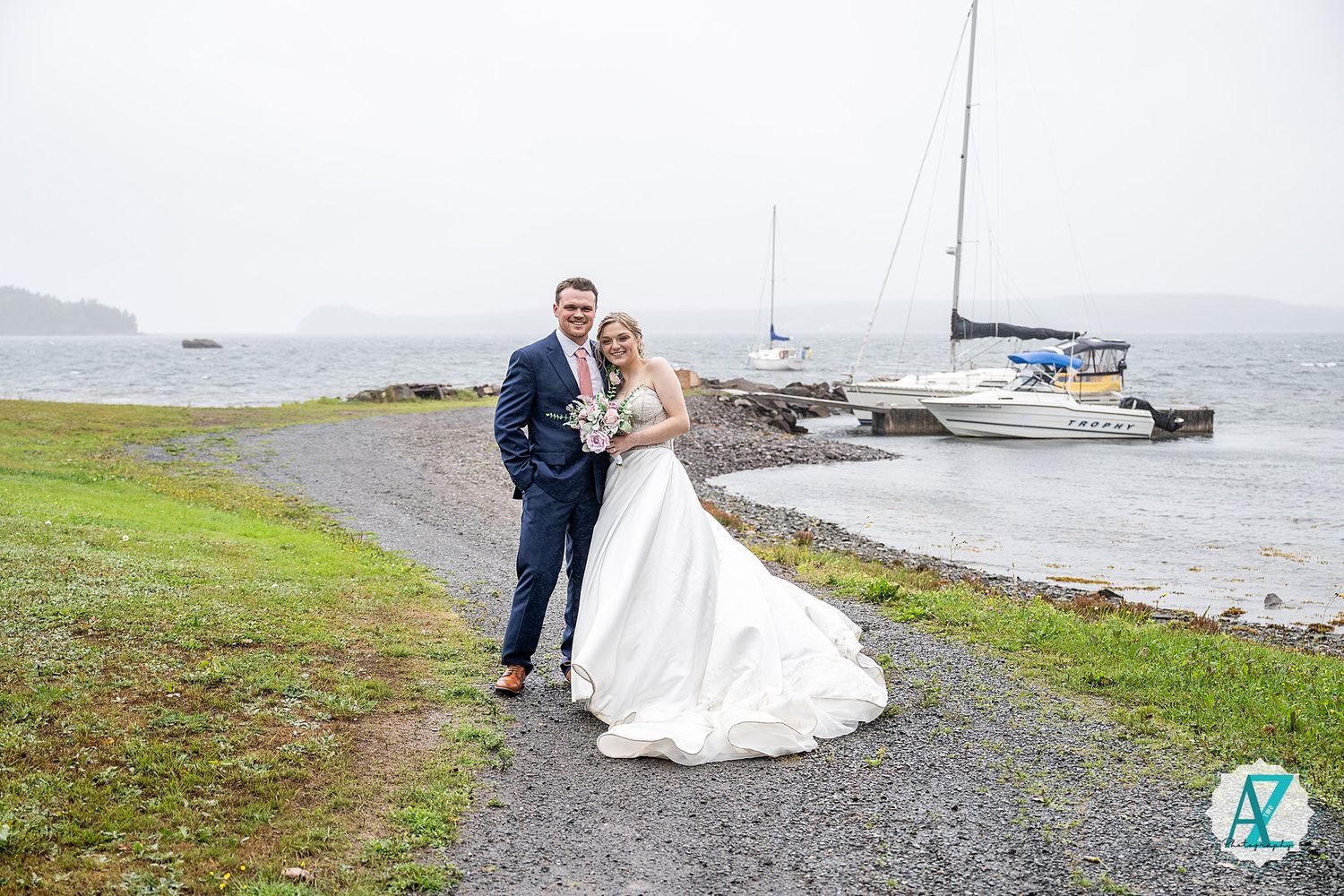 Newlyweds share romantic moments along Maine's rocky coastline with boats and water in the background.