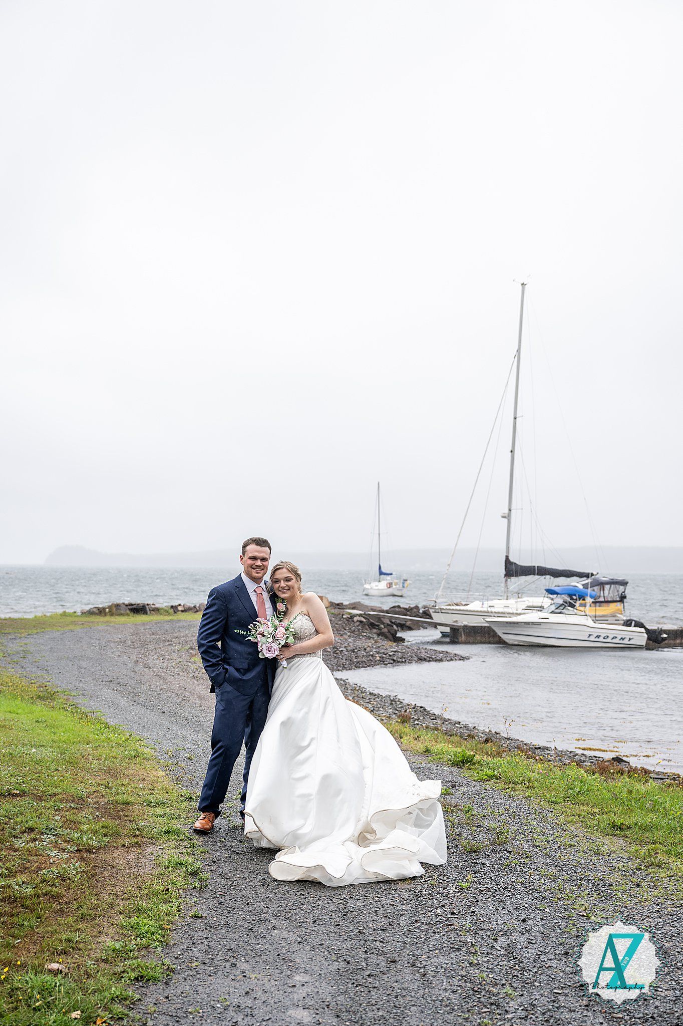 A wedding couple poses on a coastal path with sailboats and water in the background on an overcast day.