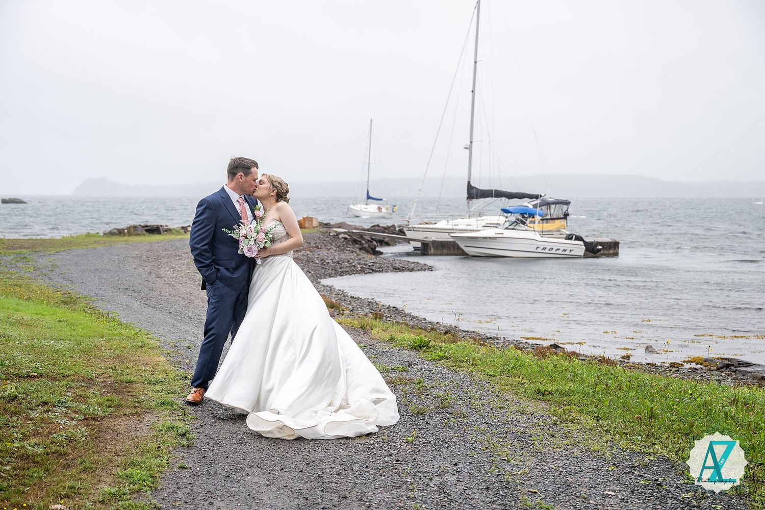 A romantic wedding couple embraces on a rocky coastal overlook with boats and ocean water in the background.