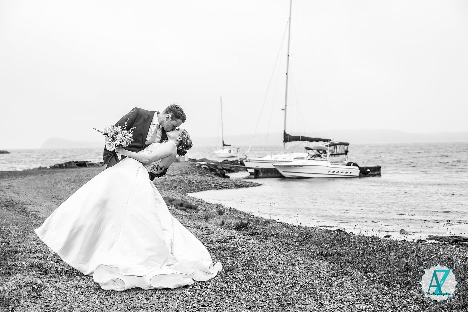 Wedding couple embracing on a misty shoreline with moored boats in the background.