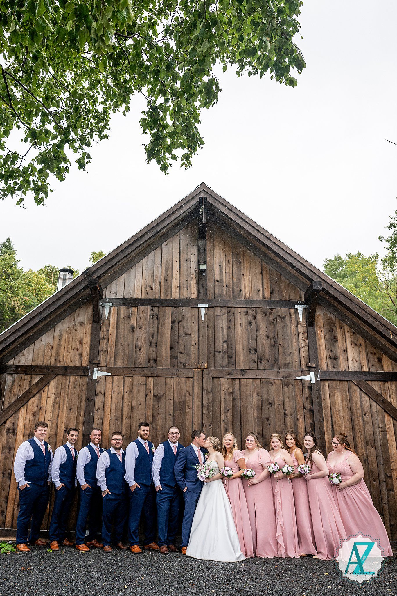 Wedding party poses in front of rustic wooden barn in blue and pink attire for group photos.