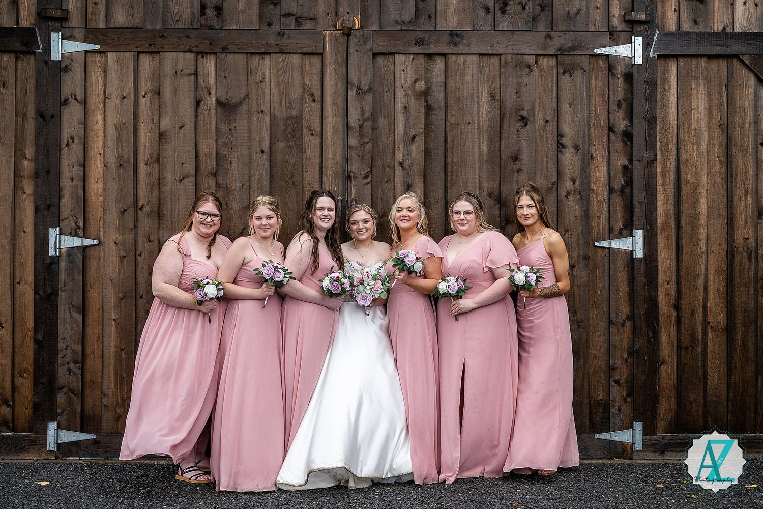 Wedding party in pink bridesmaid dresses pose in front of rustic wooden barn exterior.