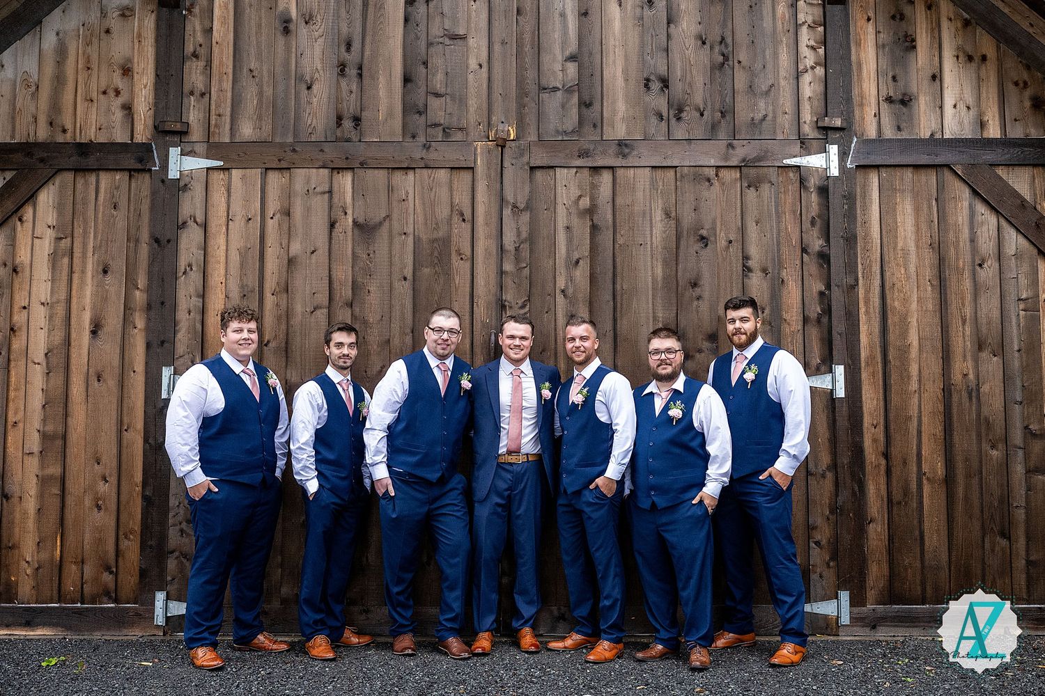 Groomsmen in navy vests and dress shirts lined up against rustic wooden barn backdrop.