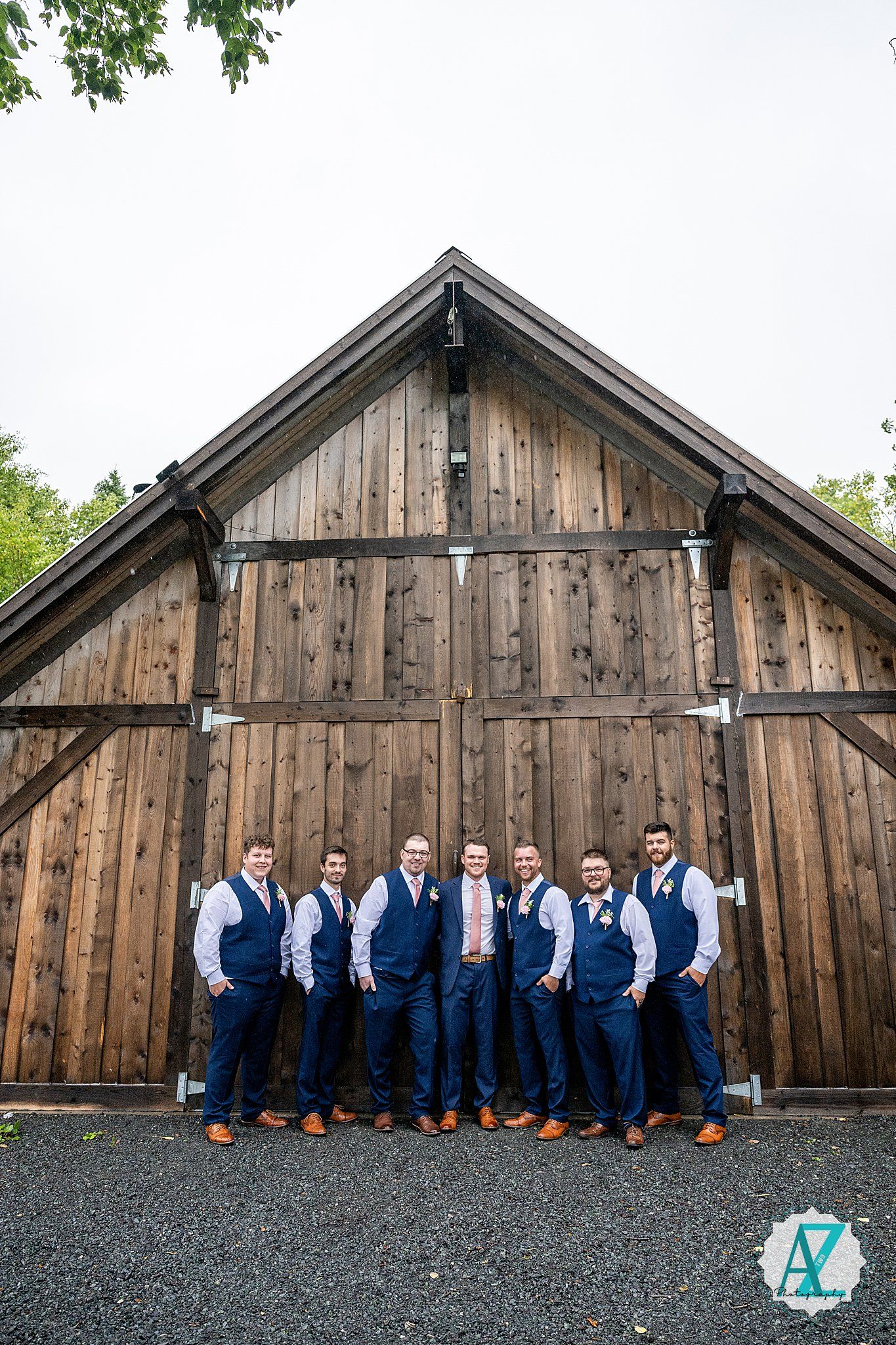 Wedding party members in coordinated navy attire pose outside a rustic wooden barn.