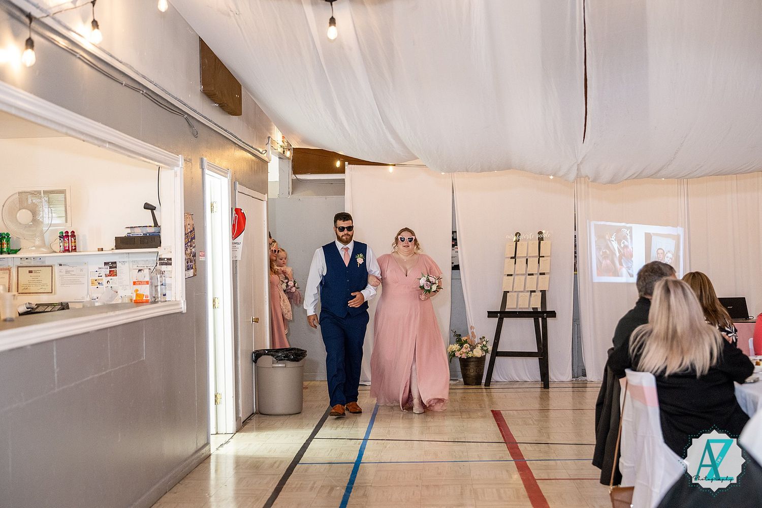 People in formal attire browse through a bright white gallery space displaying artwork on the walls at an exhibition opening.