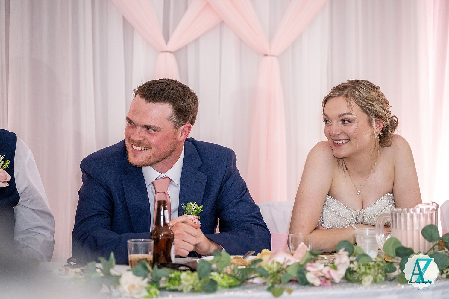 Wedding party seated at head table during reception laughing and enjoying speeches.