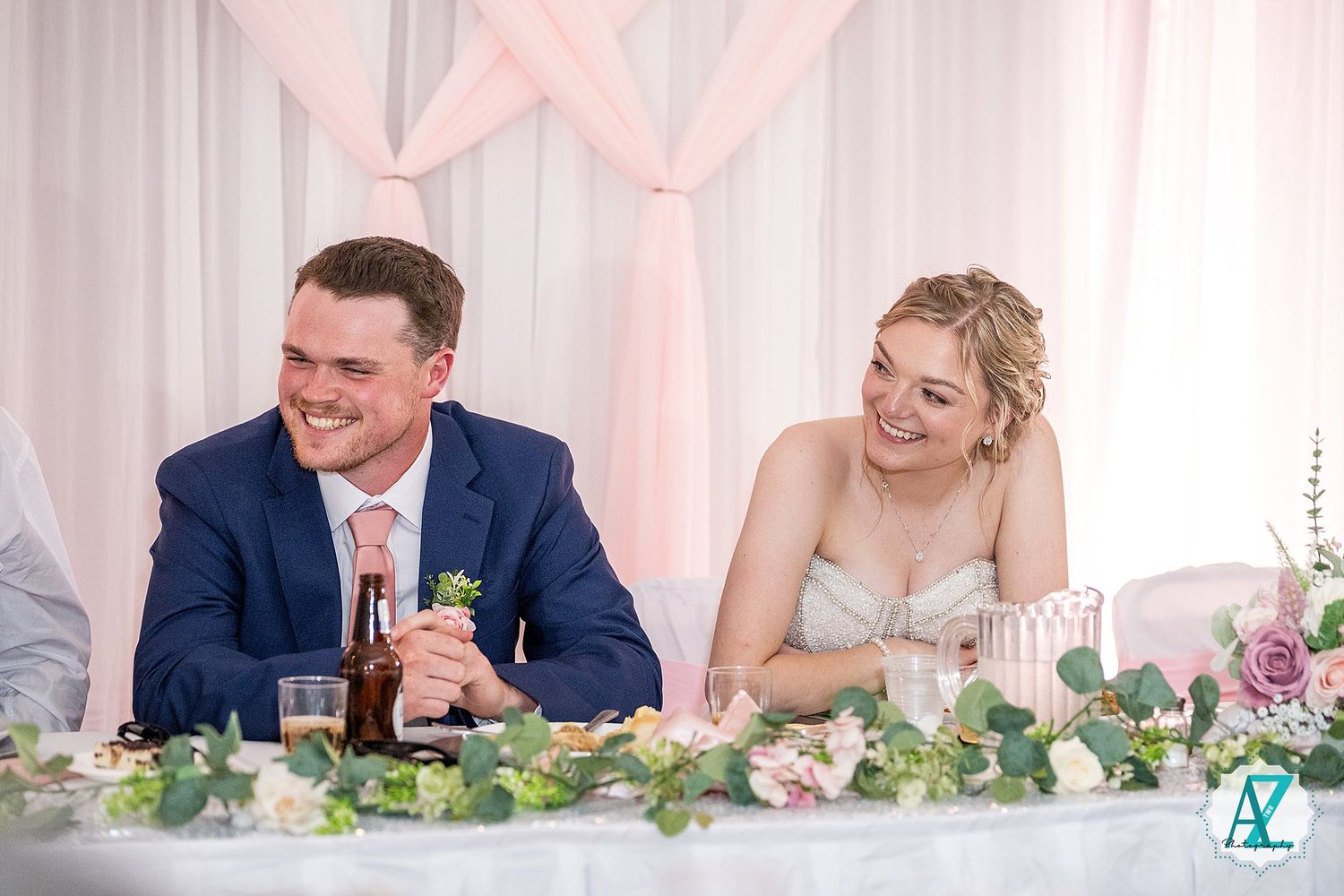 Wedding reception head table decorated with greenery garland and soft pink lighting.