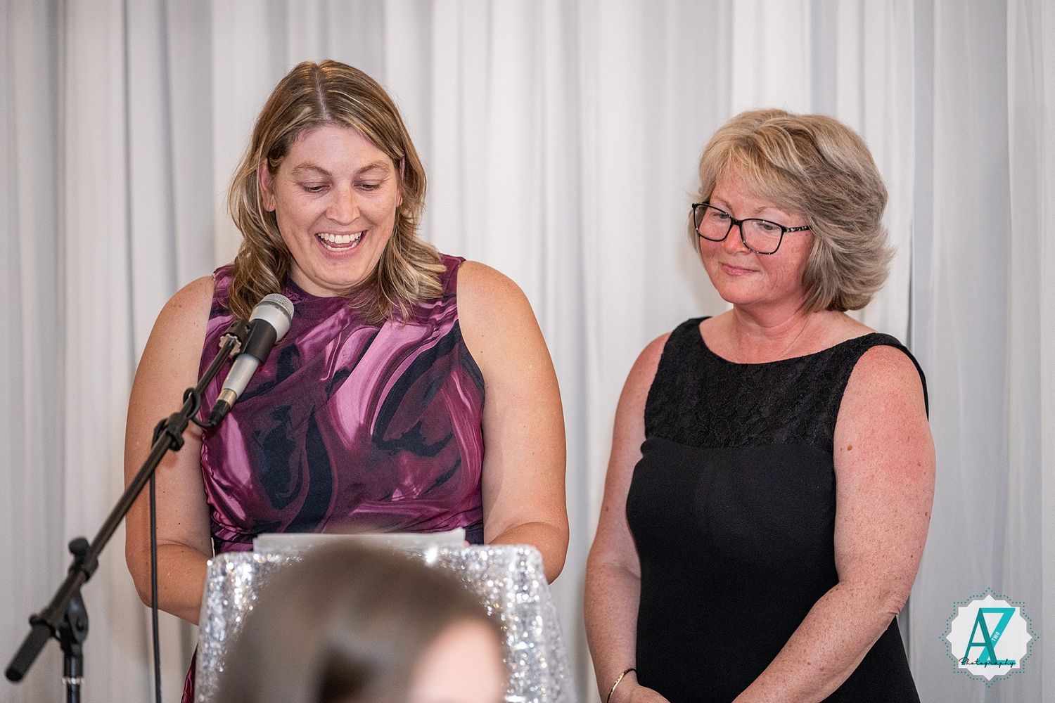 Two women at a podium share a light moment during a speaking event with one speaking into a microphone.