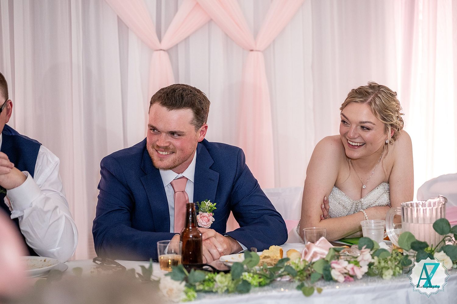 Wedding party members seated at decorated reception table with floral centerpieces and white drapes in background.