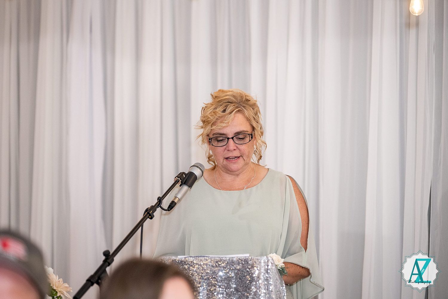 People in formal attire stand at podiums with microphones during what appears to be a wedding reception.