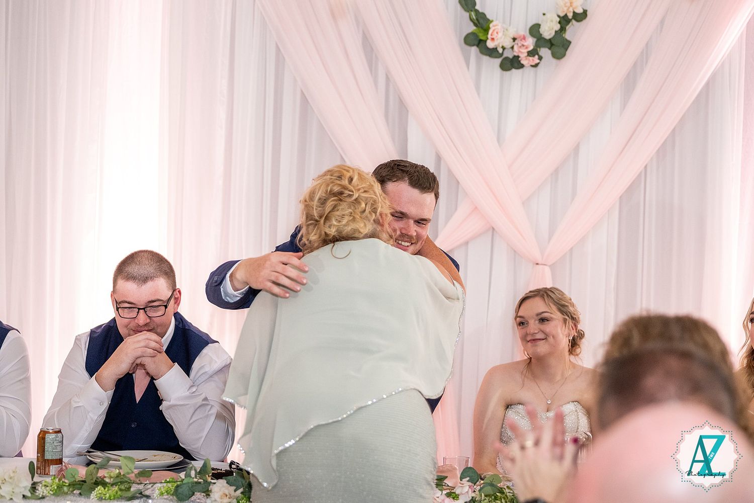 Wedding party seated at long head table with pink and white draping in background during reception.