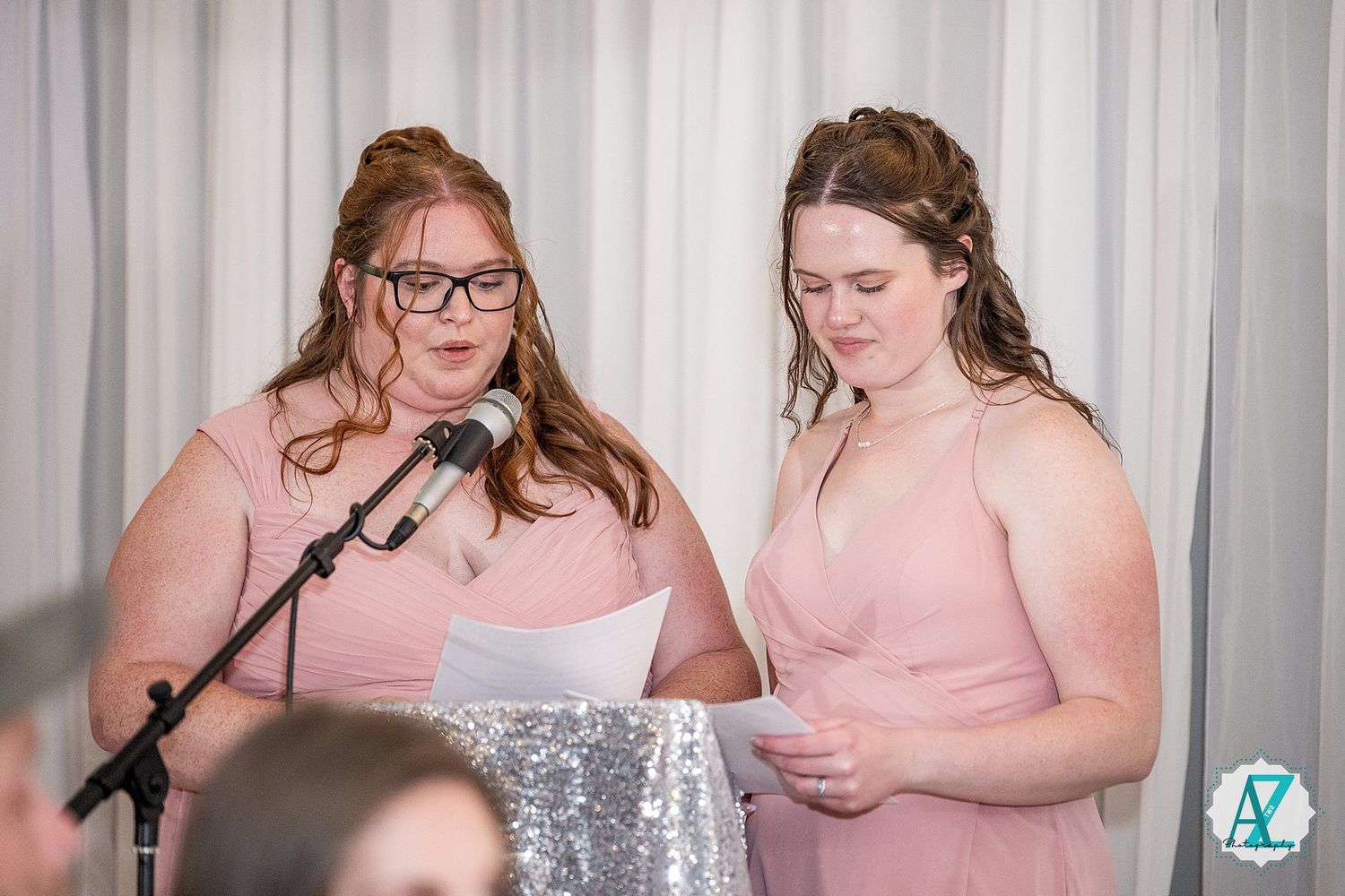 Wedding reception speakers in pink dresses stand at microphones against a white backdrop.