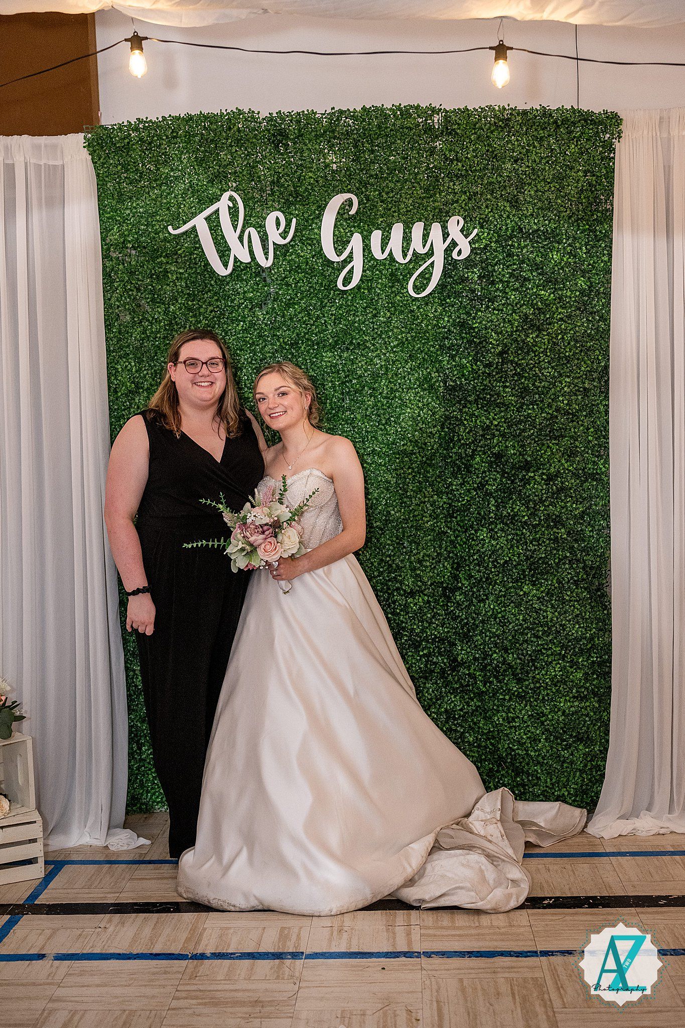 A wedding photoshoot setup with a decorative green hedge wall backdrop and white curtains in an indoor venue.