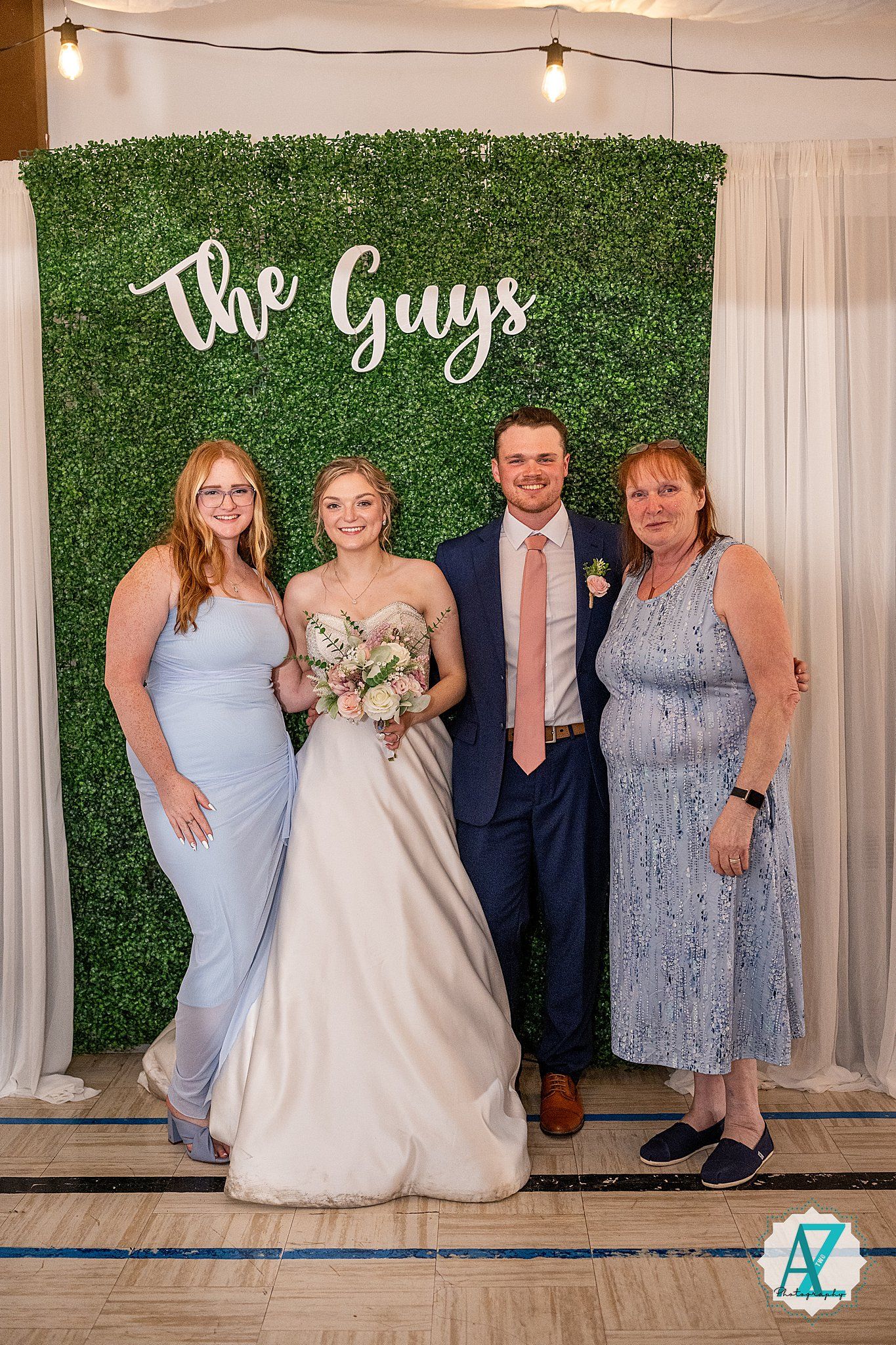 Wedding party posing against green hedge wall backdrop with white curtains and string lights at reception venue.