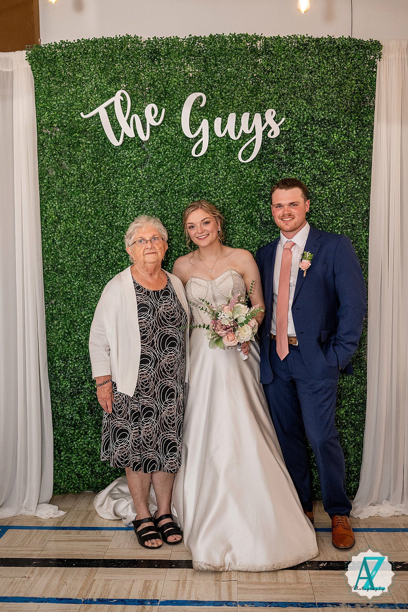 Wedding photo with guests posing against a green hedge wall with white scripted text reading "The Guys" above.
