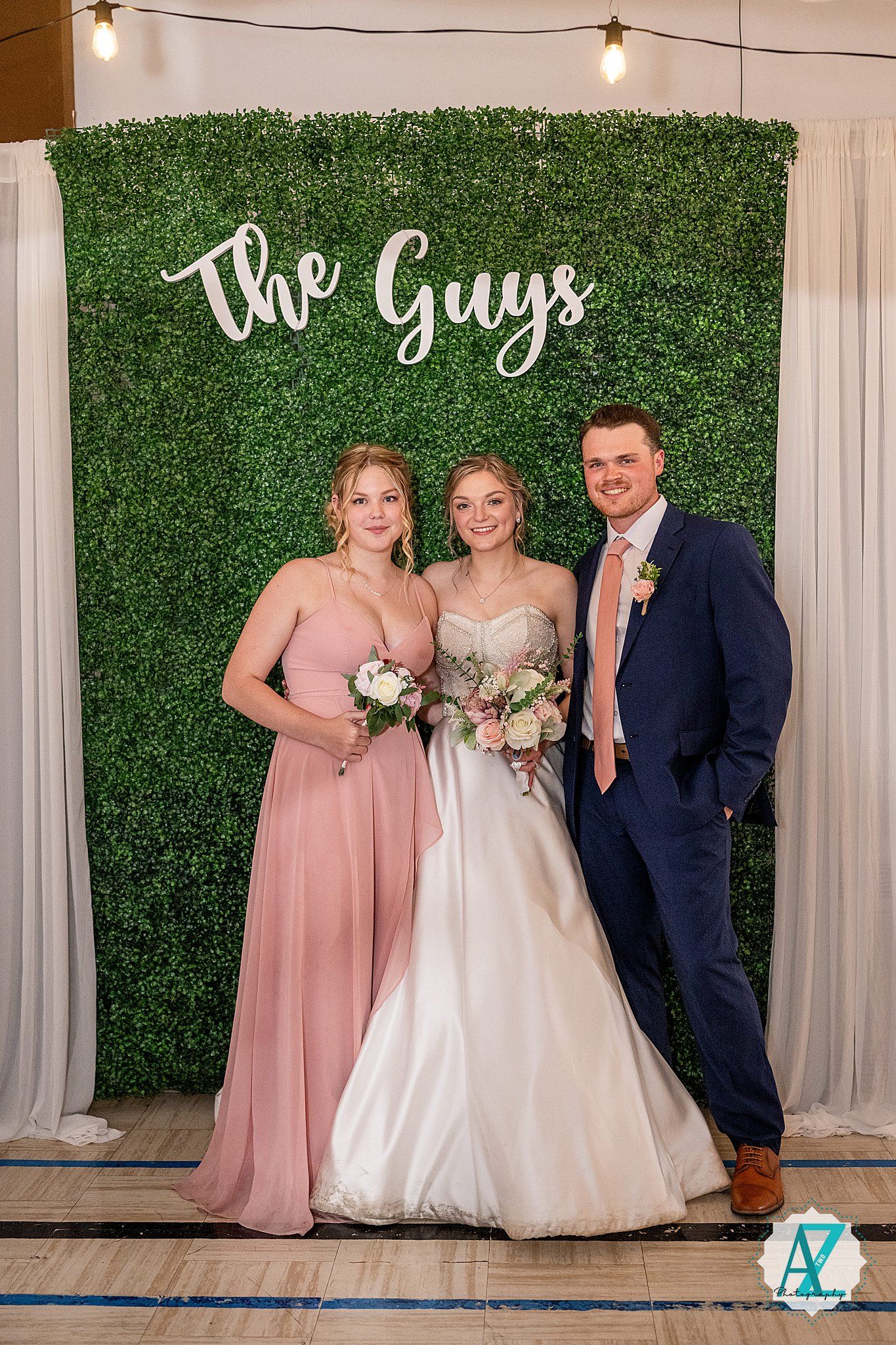 Wedding party poses against a green hedge wall backdrop with string lights and white drapes at an indoor venue.