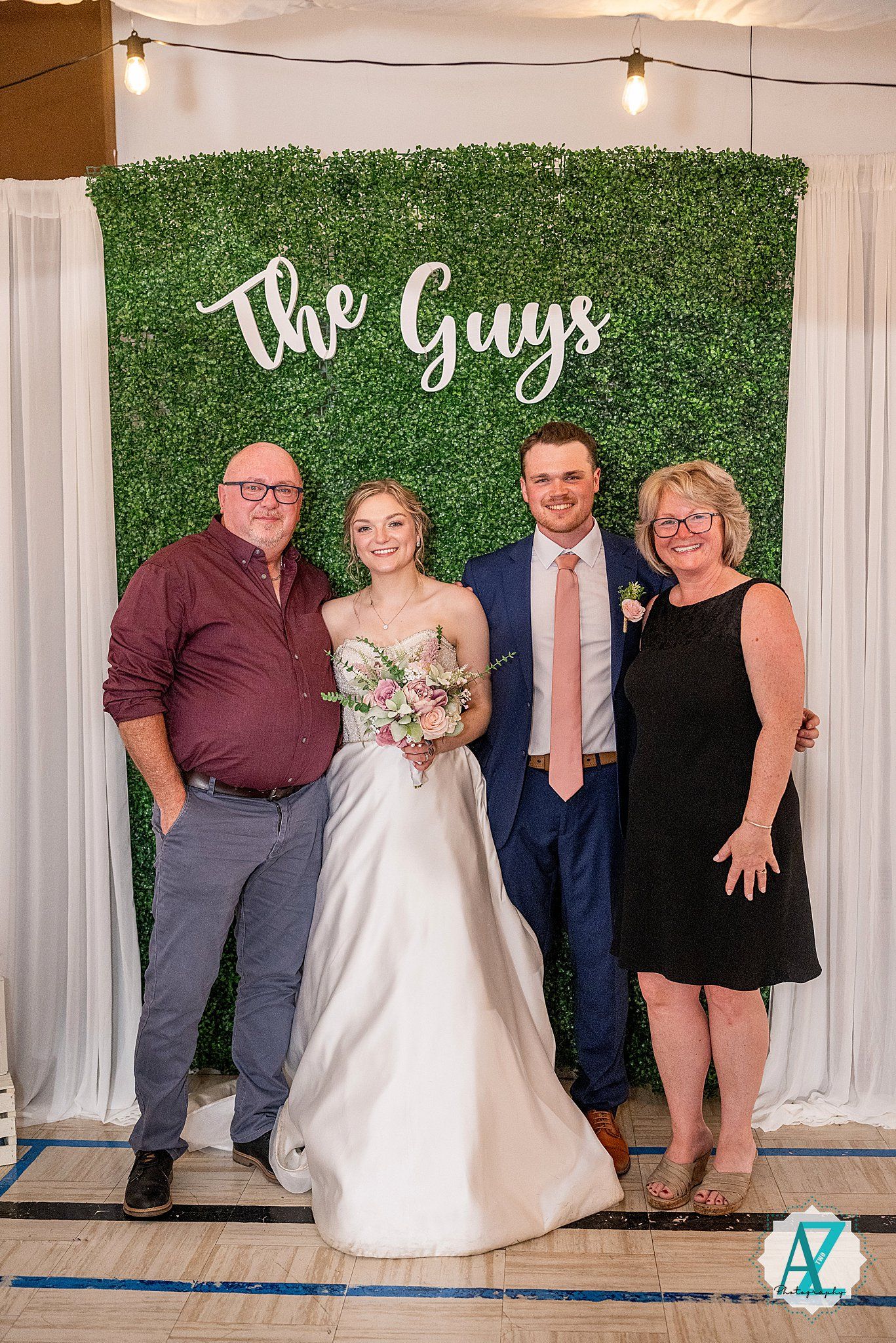 Wedding celebration group shot with decorative green wall backdrop and 'The Guys' text visible.