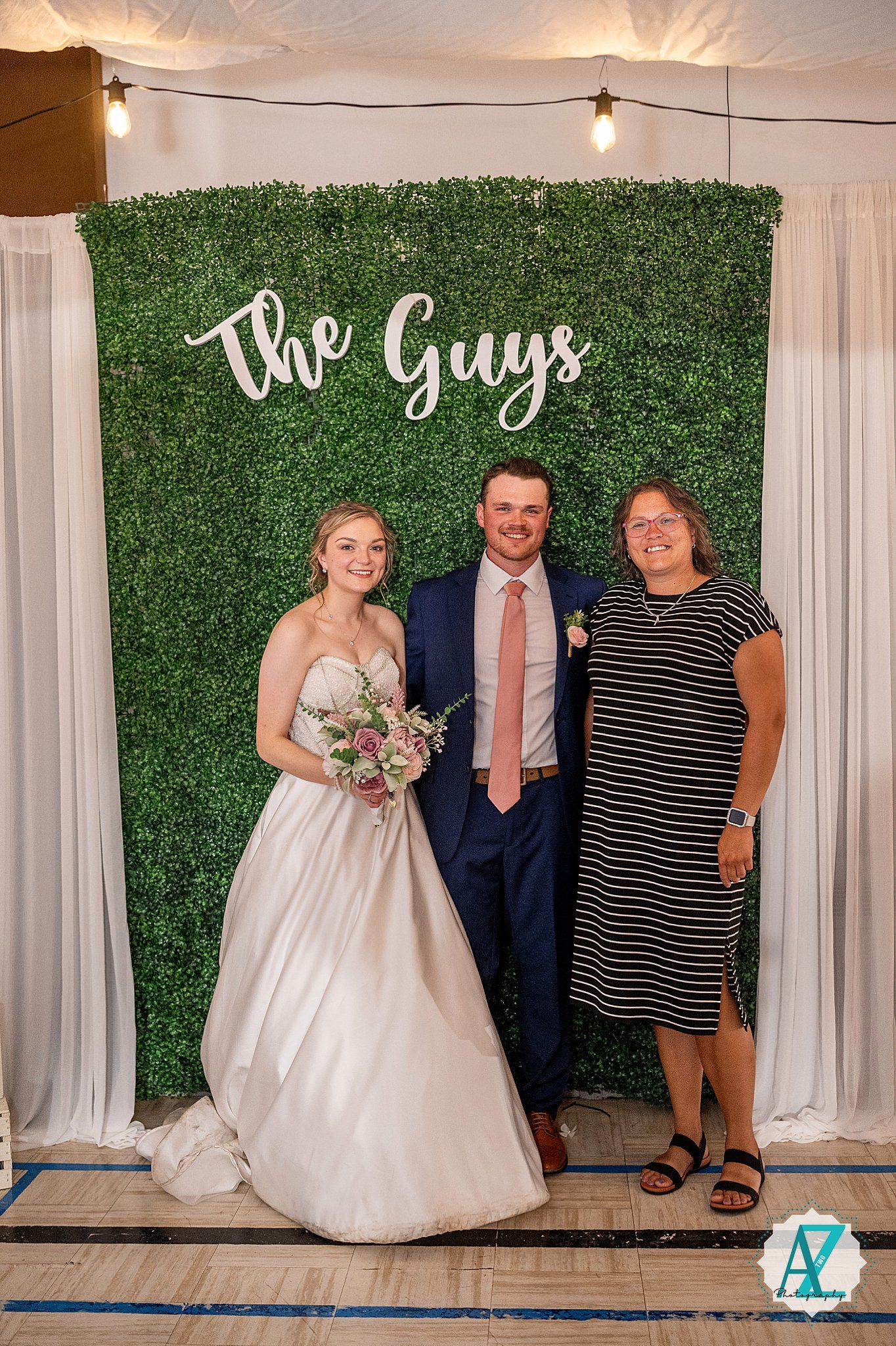 Wedding photo setup featuring decorative green hedge wall backdrop with 'The Guys' text and string lights at reception venue.