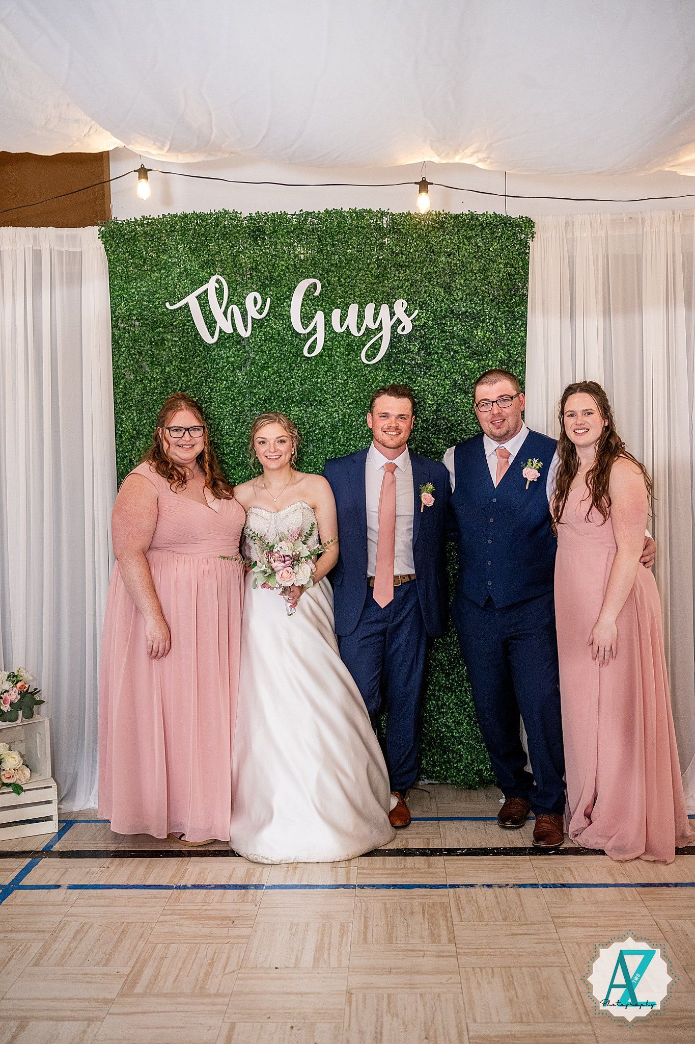 Wedding party poses in alternating pink dresses and navy suits against a white backdrop with green hedge wall.