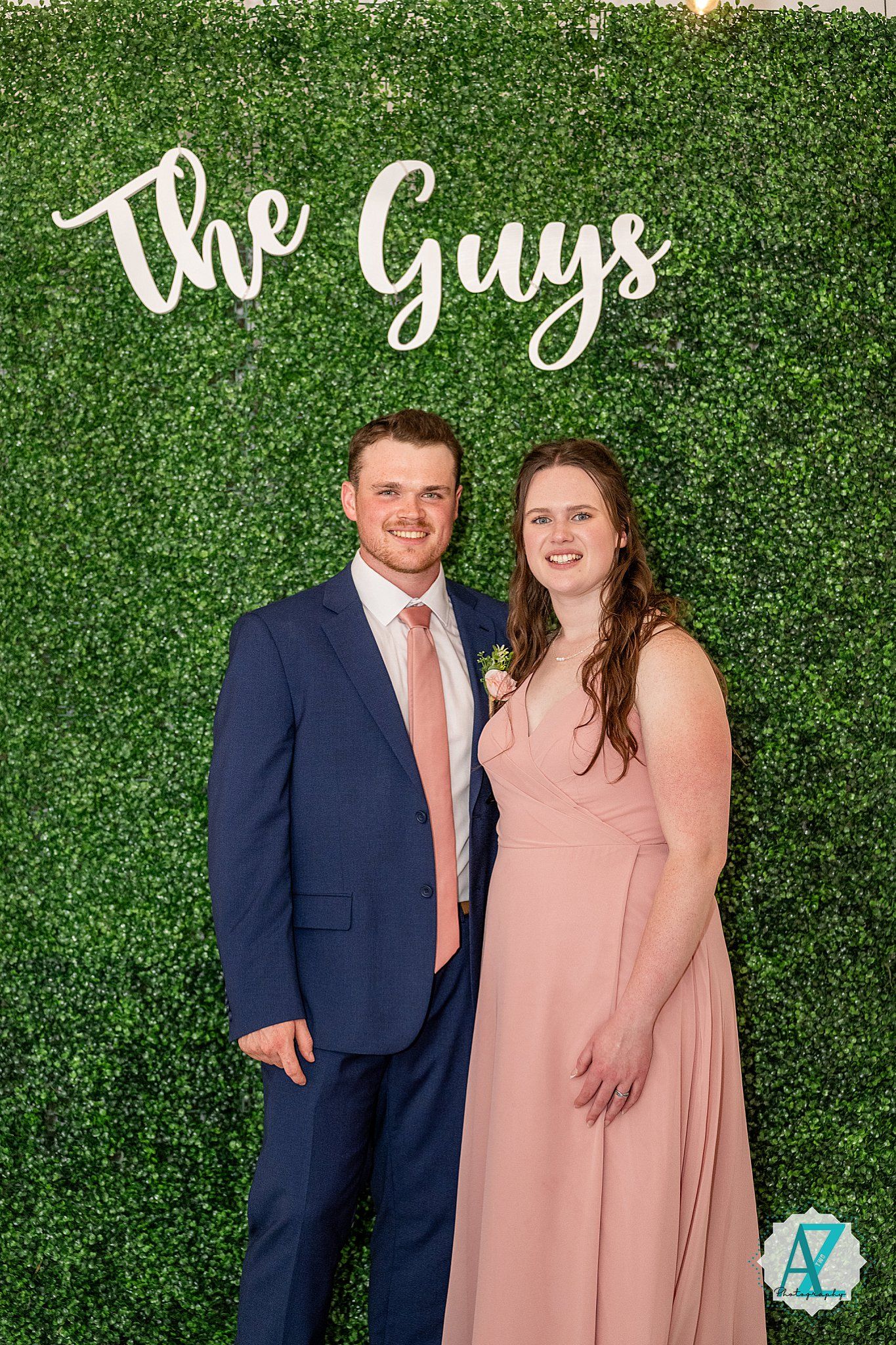A couple poses in front of a green hedge backdrop wearing coral and navy formal attire.