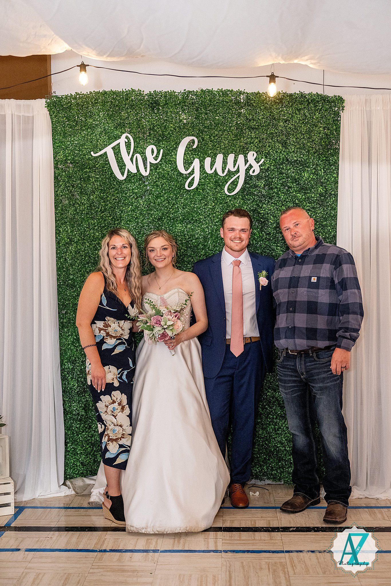 Wedding party stands in front of green hedge backdrop with white curtains at indoor reception venue.