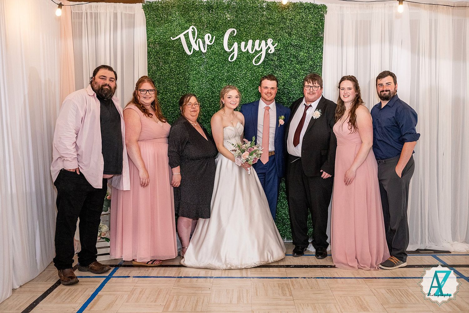 Wedding celebration group photo with decorative green wall and white drapery background.