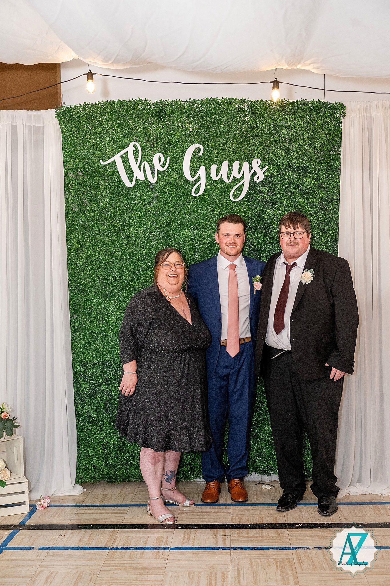 Wedding photo setup with green hedge wall backdrop displaying 'The Guys' text and white curtain decorations.