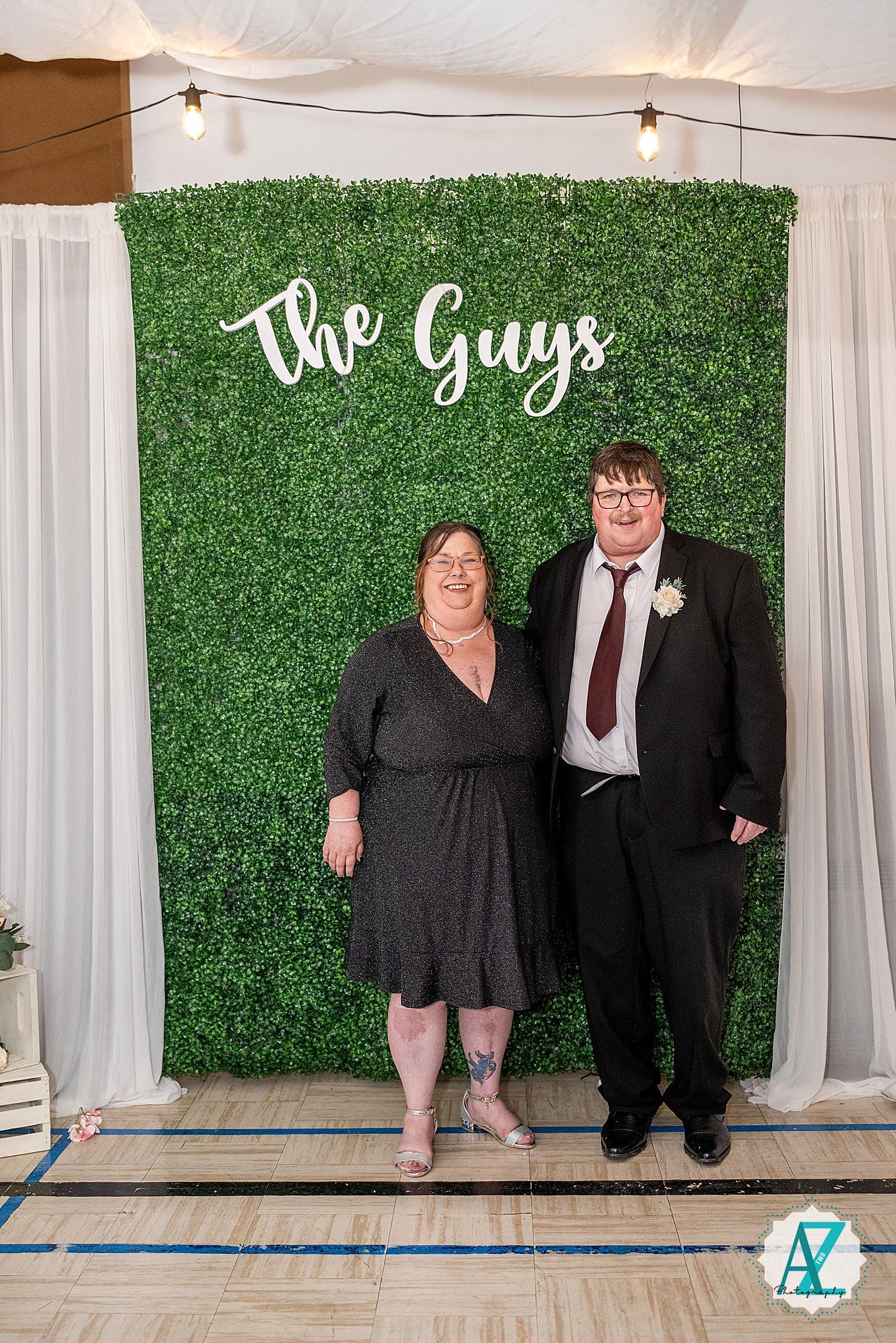 Wedding guests pose in front of a green hedge wall with white curtains and string lights at an indoor venue.