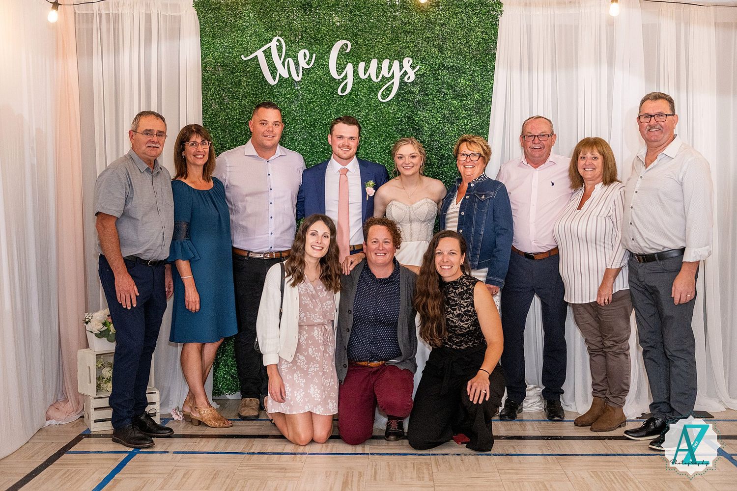 Group photo taken at a wedding event with people lined up against a white curtain backdrop and rustic decor.