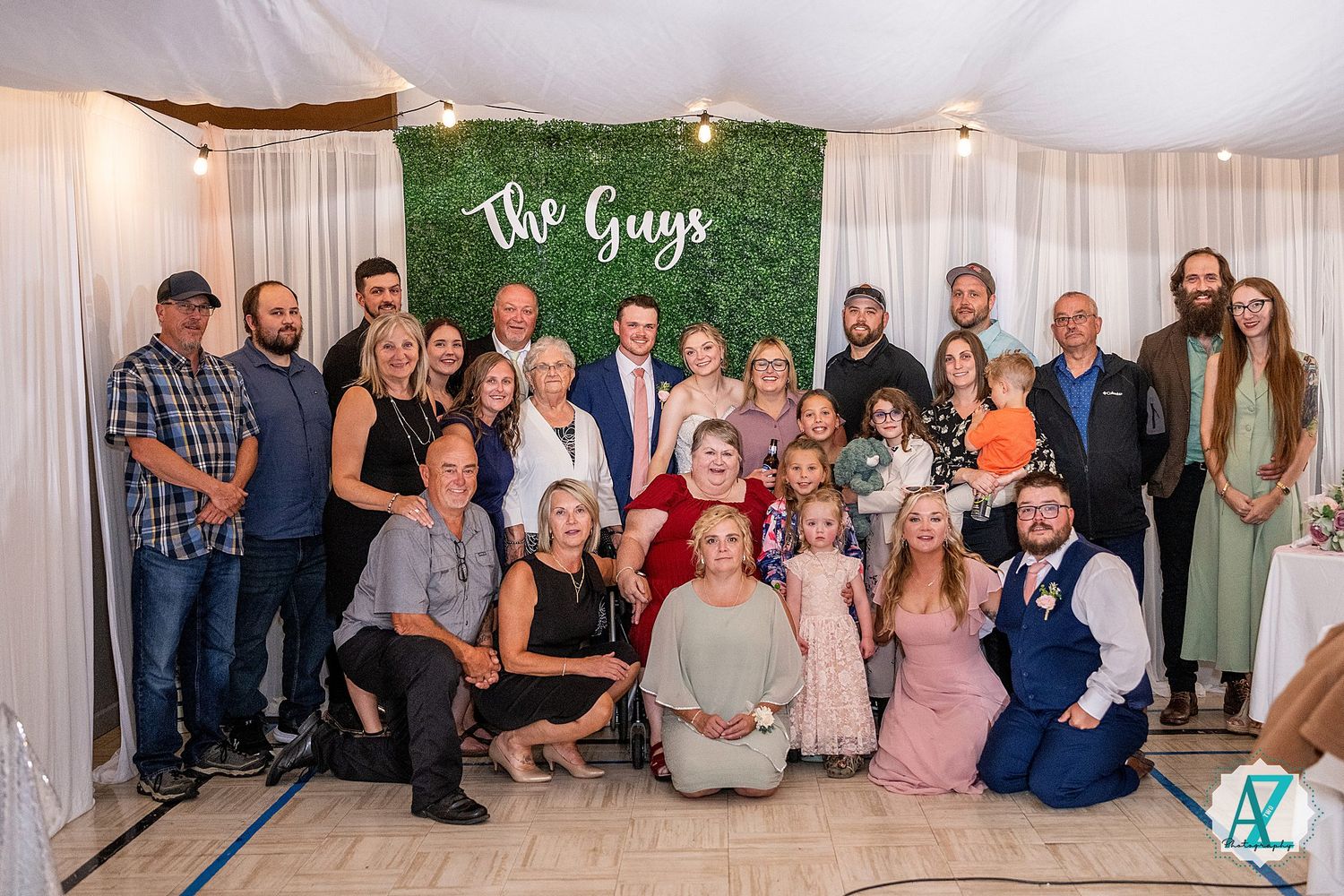 Wedding party group photo in front of decorative green wall and white curtain backdrop at reception venue.