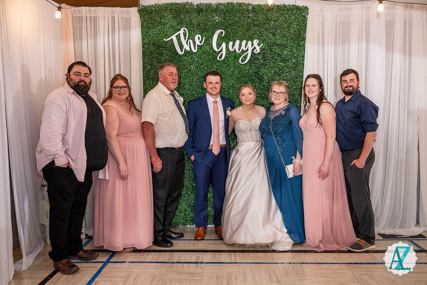Wedding party members pose together against a white backdrop with decorative wall signage and fabric draping.