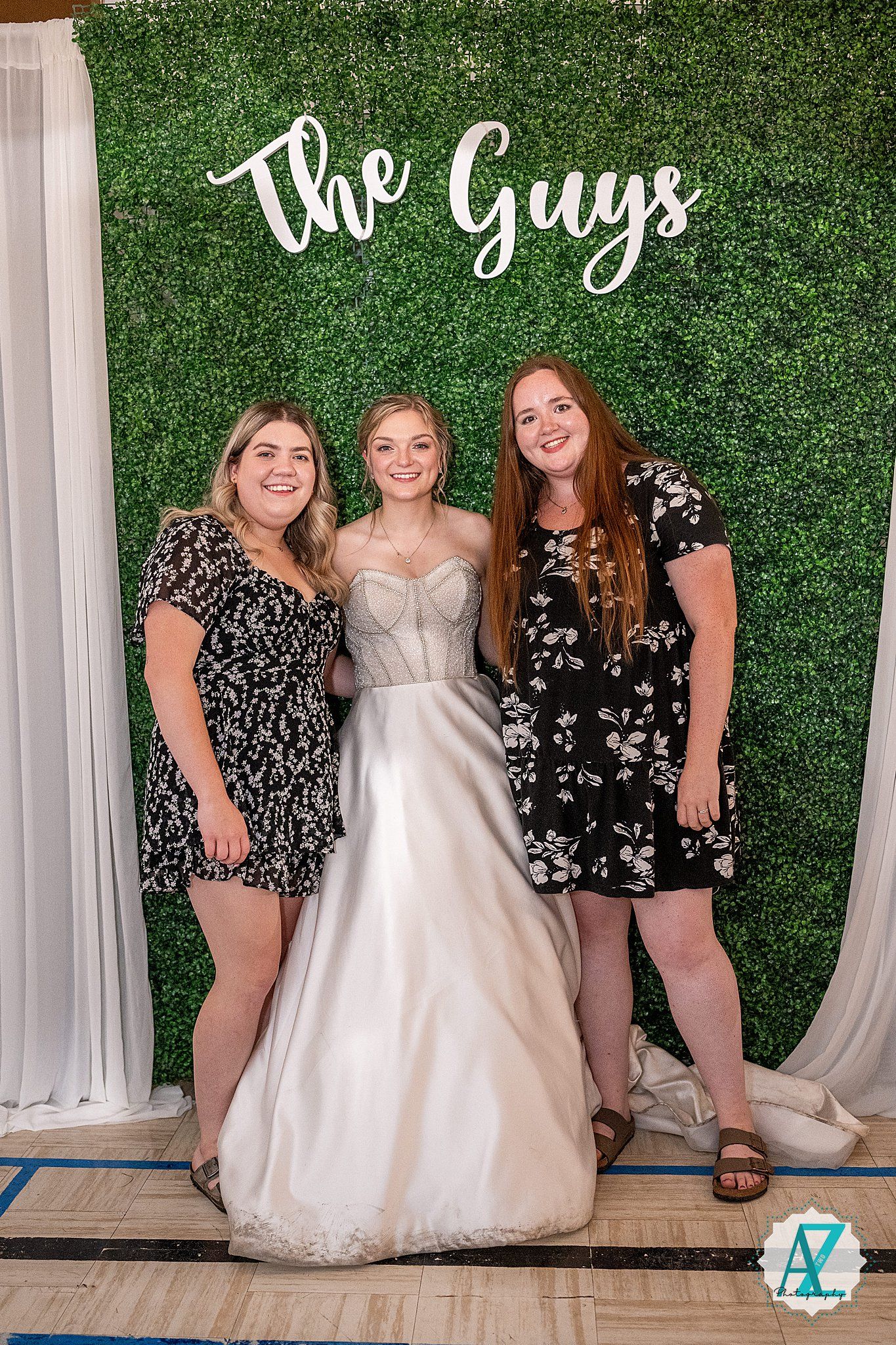 Group photo of bridesmaids in dark dresses posing with bride in white gown against green wall backdrop.