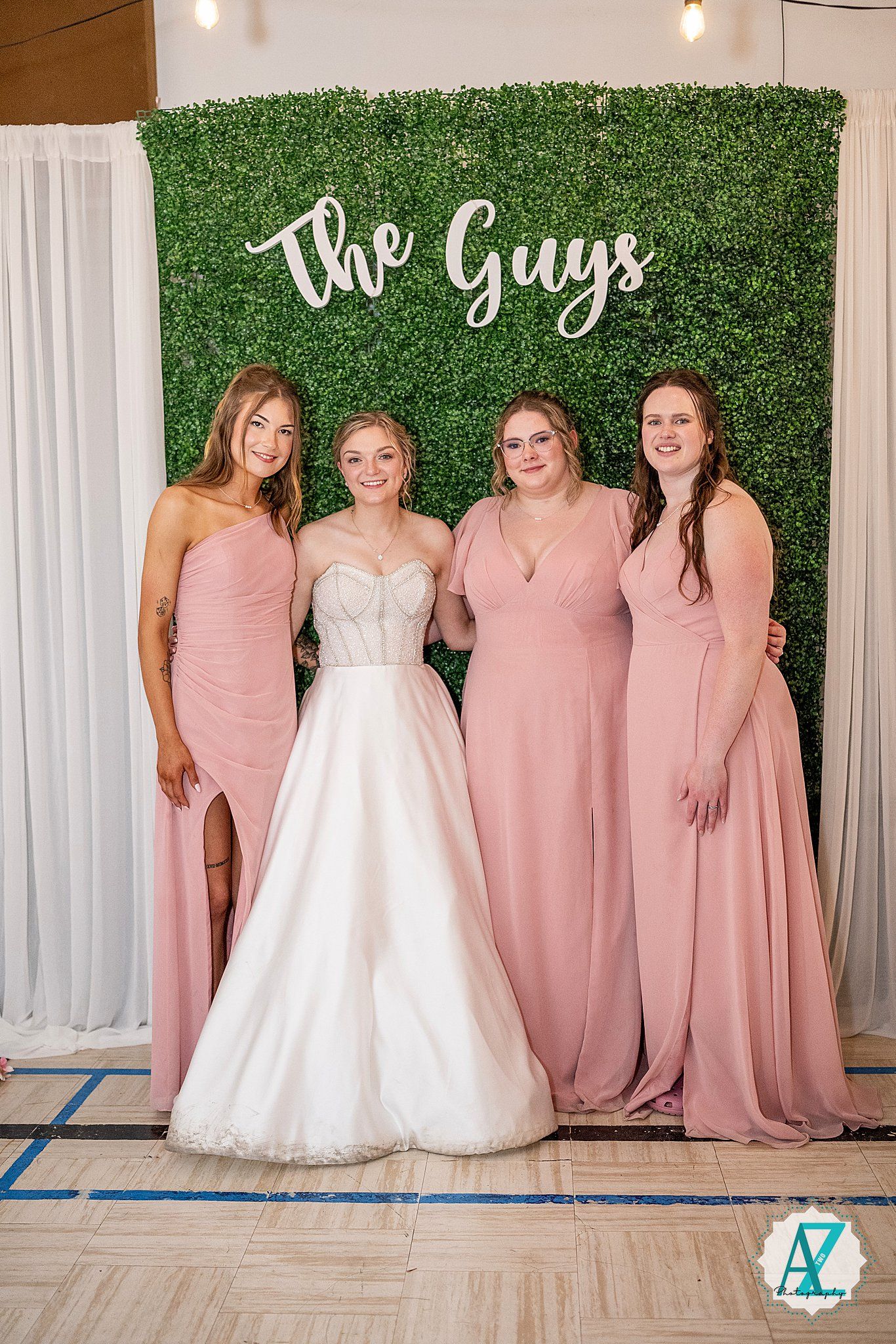 Wedding party poses in pink bridesmaid dresses and white bridal gowns against a green hedge backdrop.
