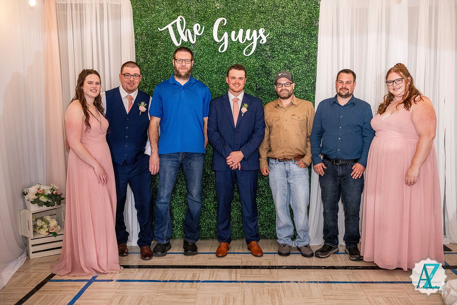 Wedding party group poses in front of a greenery wall backdrop with white draping and decorative lights.