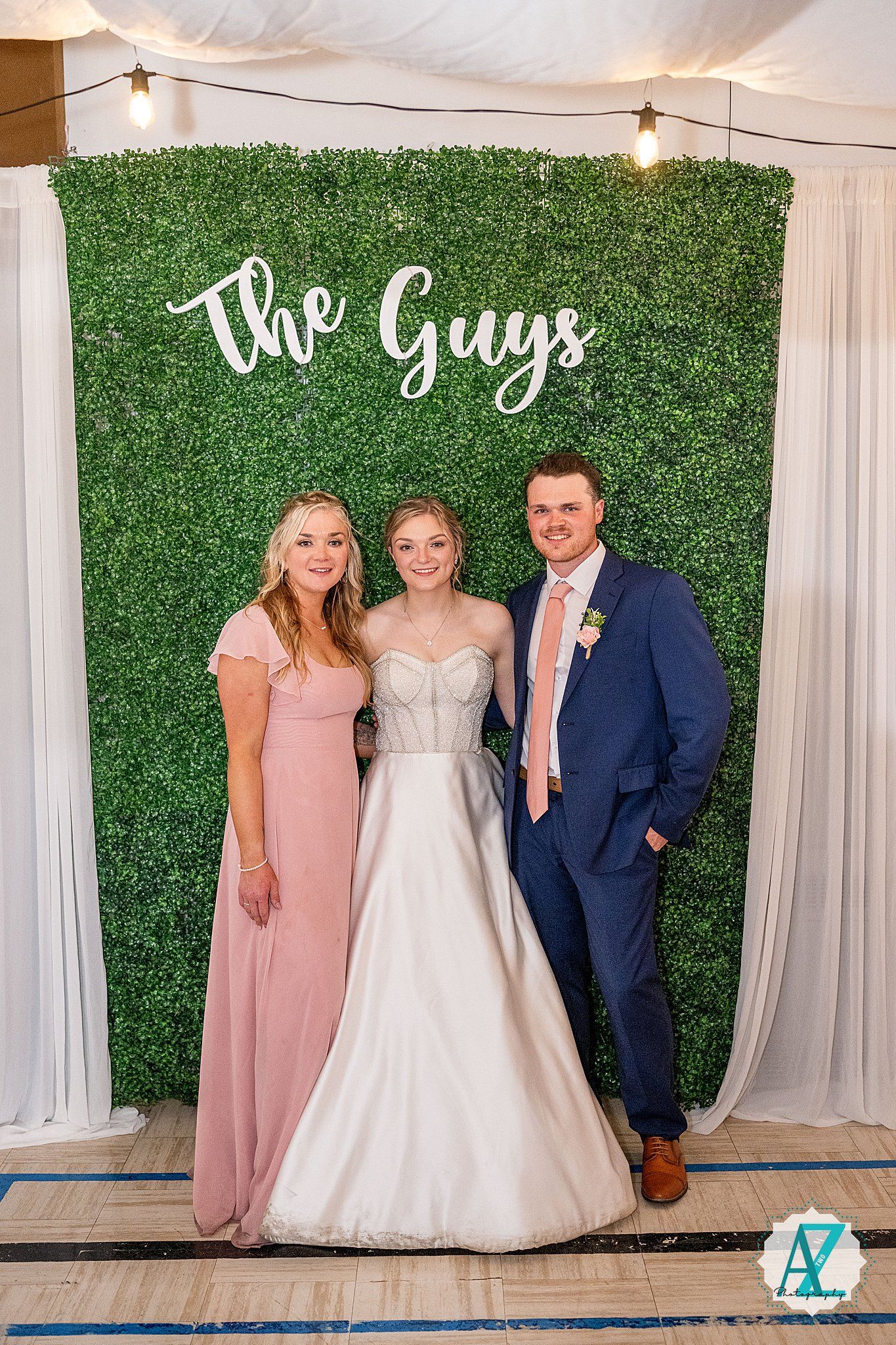 Wedding party poses against green hedge backdrop with white drapes and soft lighting at indoor venue.