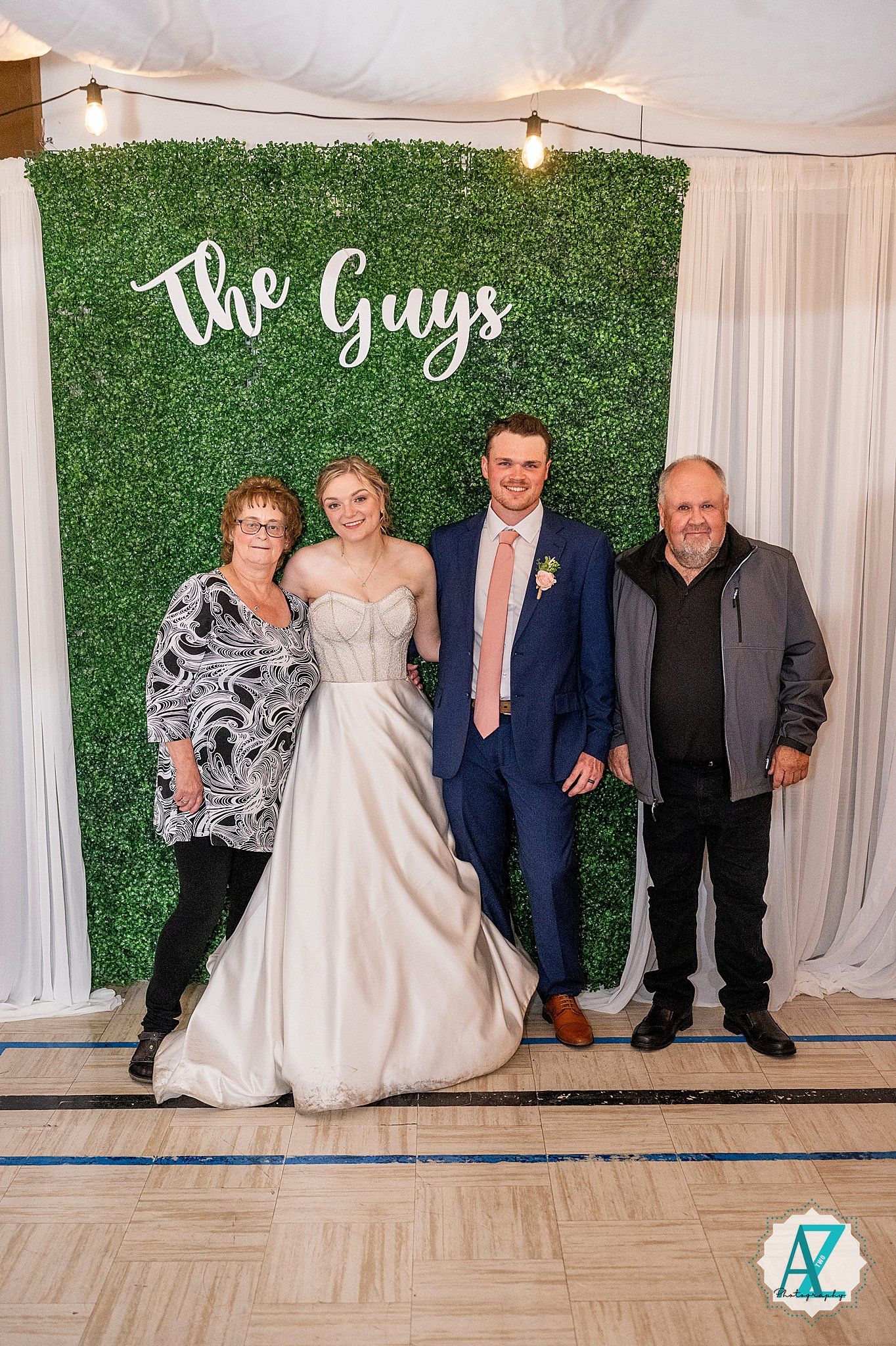Wedding group photos taken against a decorative green hedge wall with The Guys text sign and white curtains.