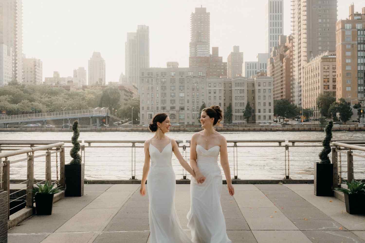 Two brides in white dresses pose for romantic wedding photos along a city river at the Sanctuary Roosevelt Island NYC