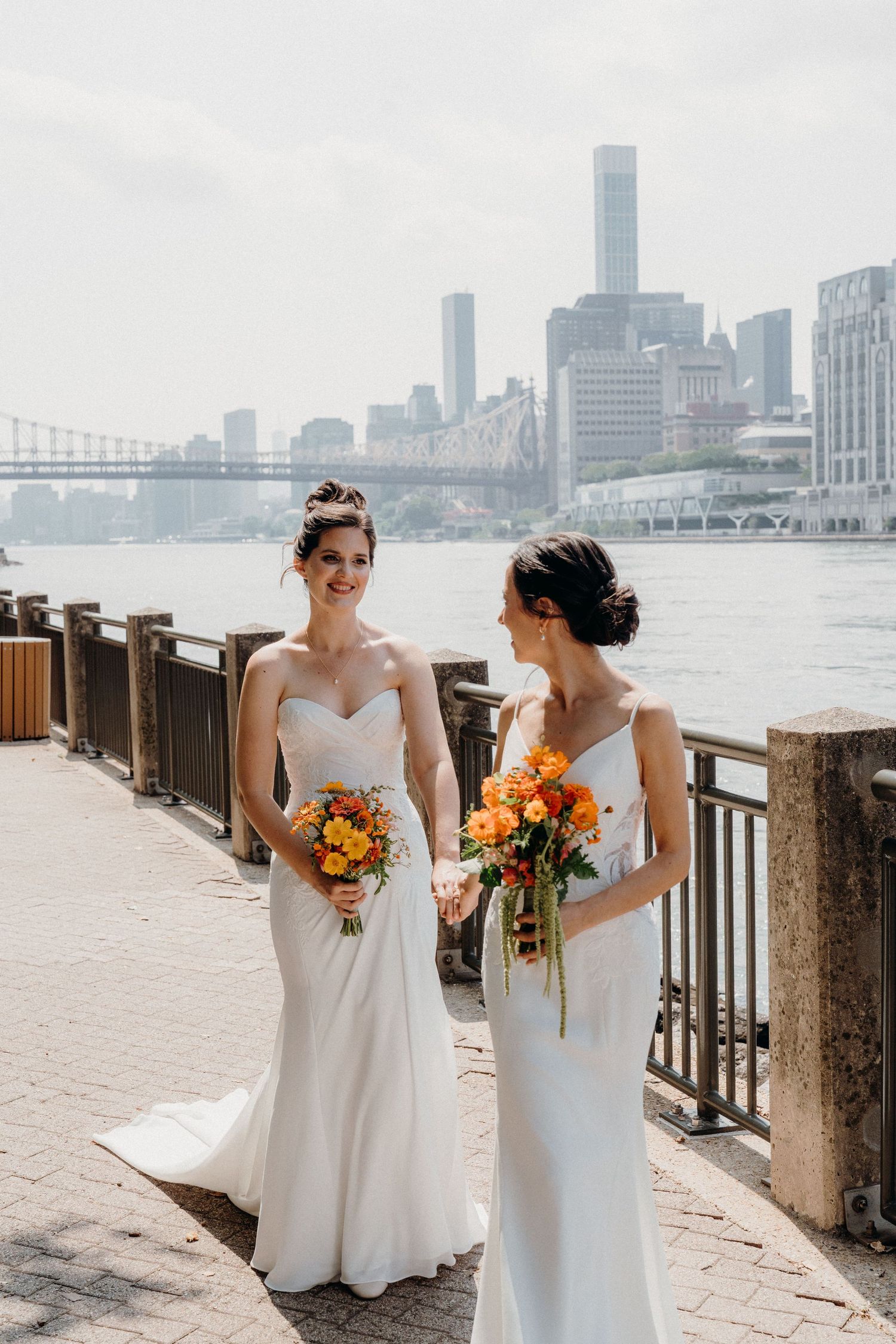 Two people in white wedding dresses pose together at the waterfront with the New York City skyline in the background.