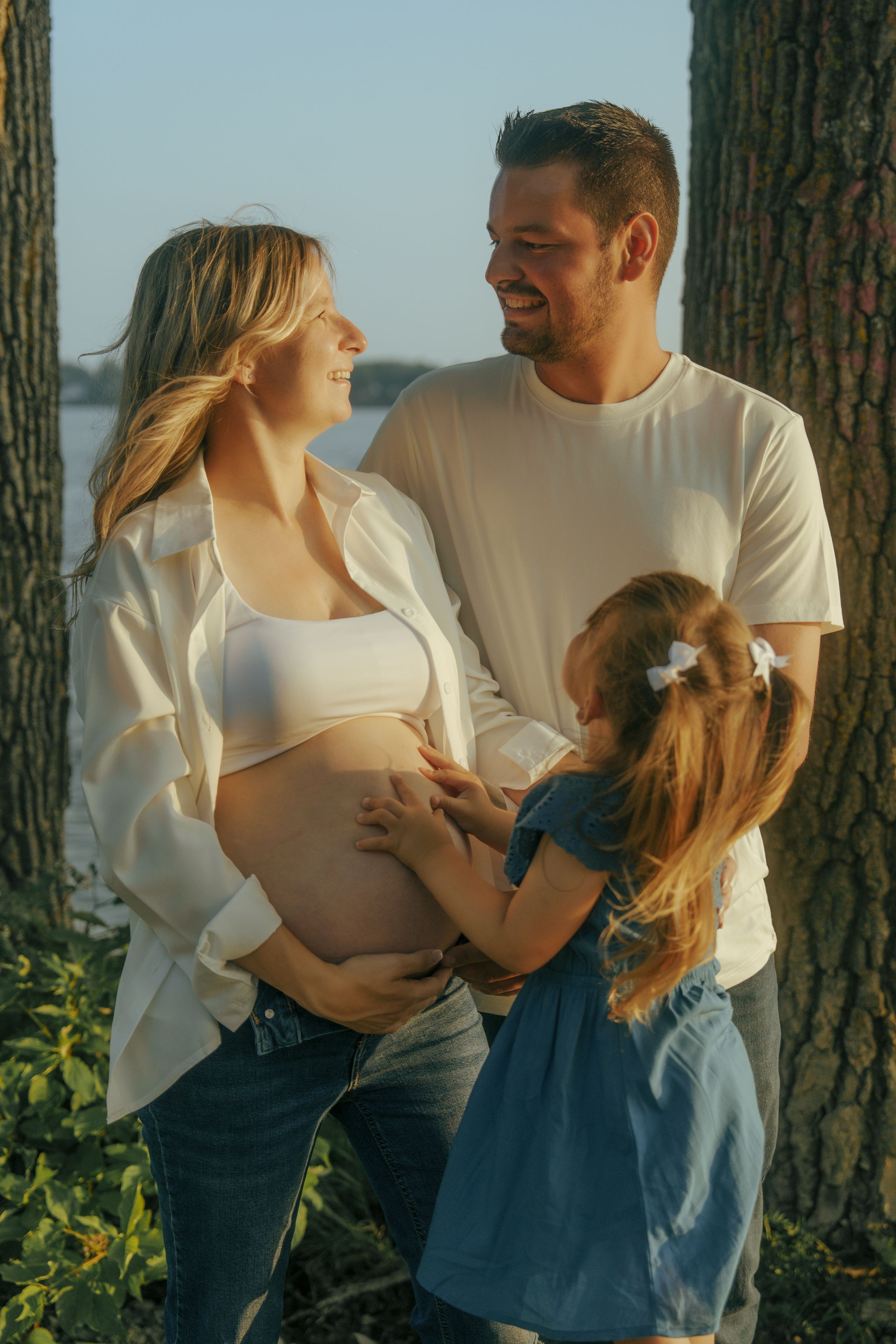 A couple in white shirts and jeans embraces tenderly against a sunlit forest backdrop.