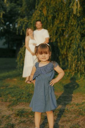 A small child in a blue dress plays under a weeping willow tree during golden hour lighting.
