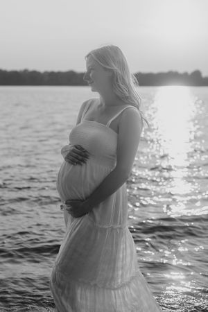 Maternity photoshoot in white dress at sunset by the water with sunlight sparkling on gentle waves.