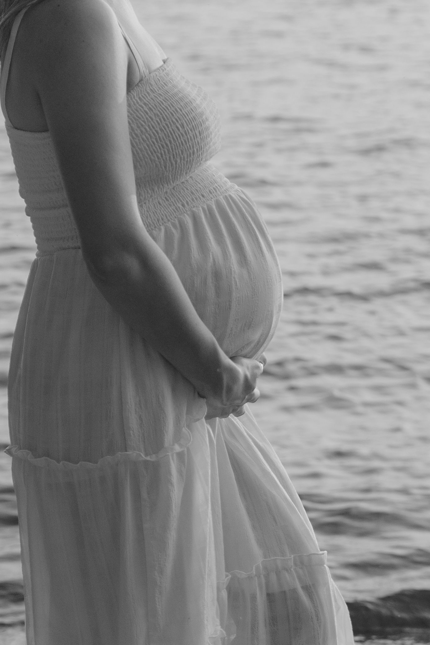 A series of black and white maternity photos showing a pregnant woman in a white dress by the water.