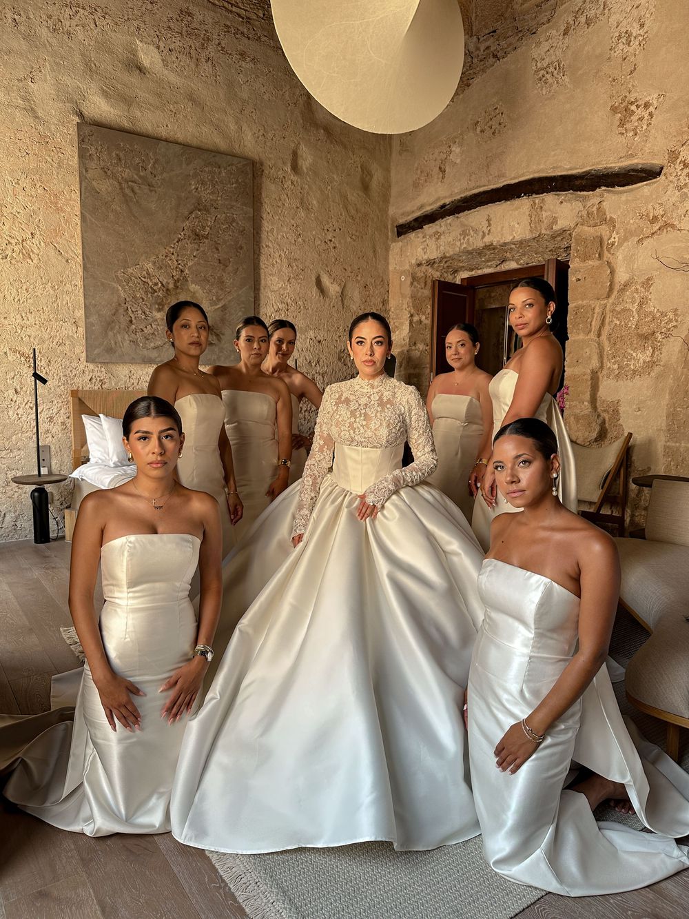 A bridal party poses in an elegant stone room wearing matching white gowns with the bride in a detailed ballgown centerpiece.