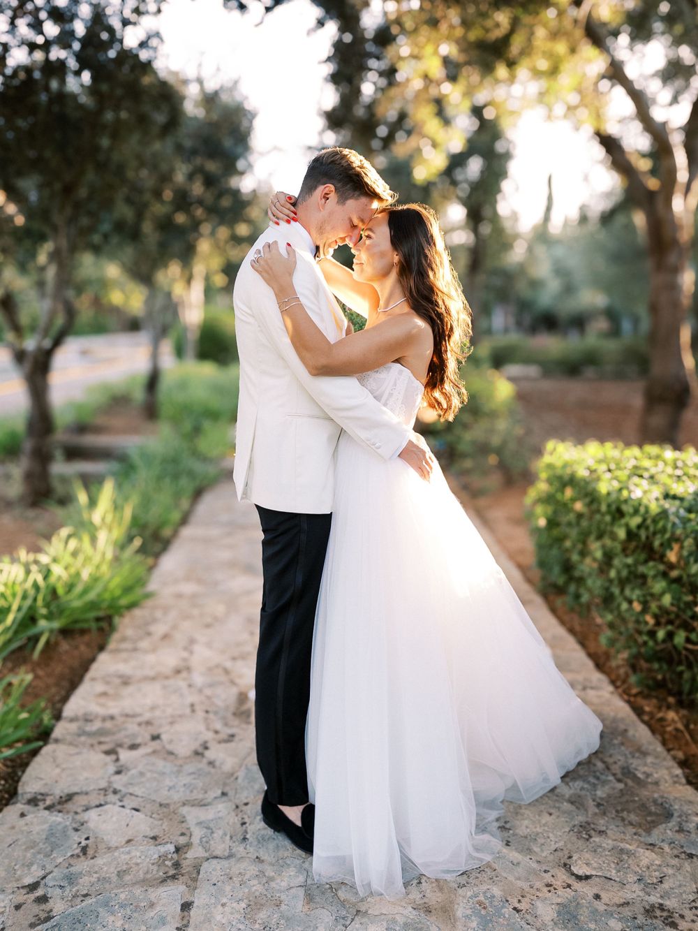 A romantic wedding couple embraces on a stone path surrounded by lush garden greenery at sunset.