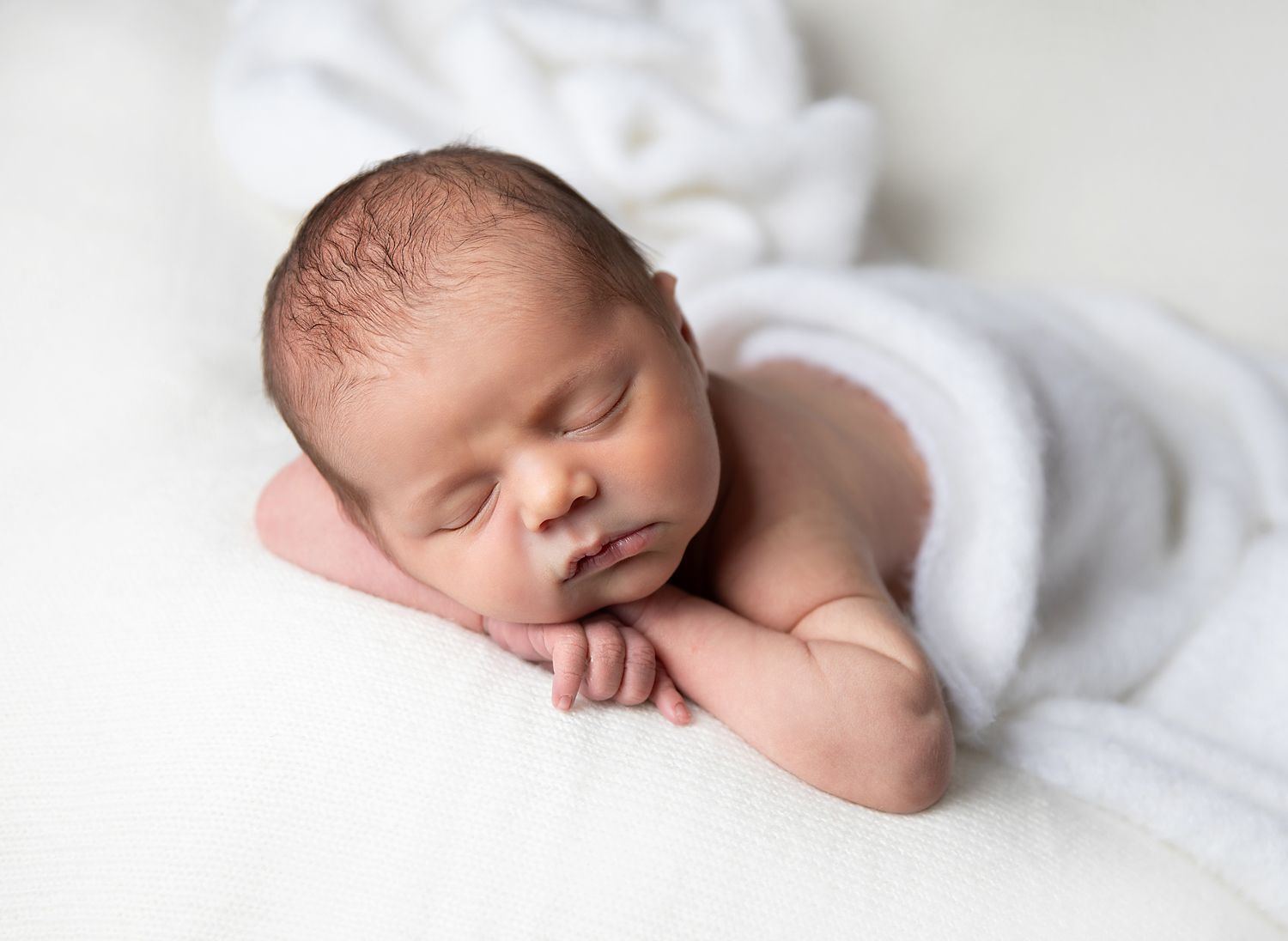 Peaceful sleeping newborn baby rests on soft white blanket with hand tucked under chin. Duding a newborn photoshoot in Sidcup